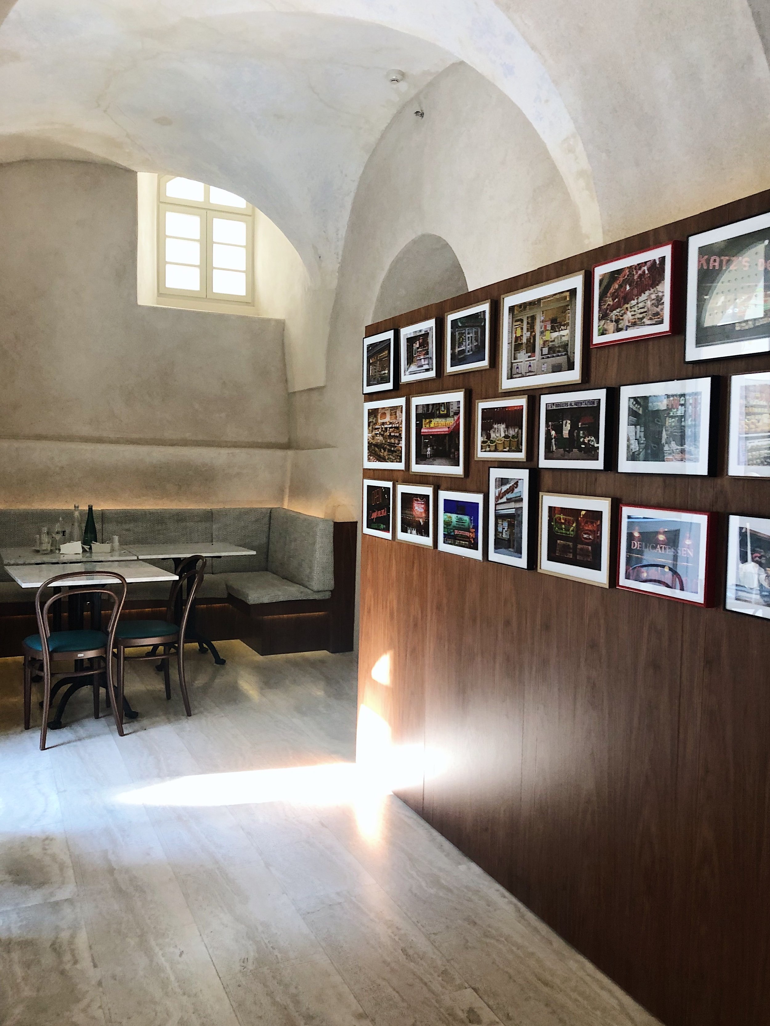 Interior of a restaurant with a small table, chairs, corner booth, and photo frames on a wooden partition wall, with a window letting in natural light.