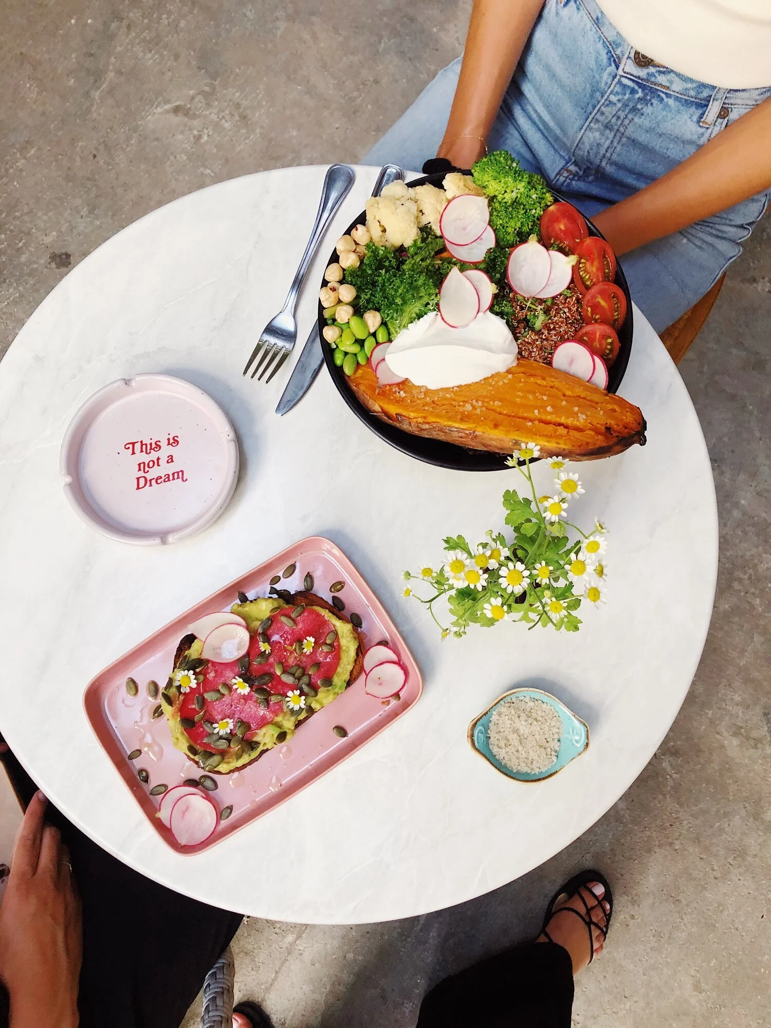 A table with a colorful salad, a bowl of rice, a small dish of dip, and a tray of avocado toast topped with radish slices, pumpkin seeds, and edible flowers. Two people are nearby, one sitting and one standing.
