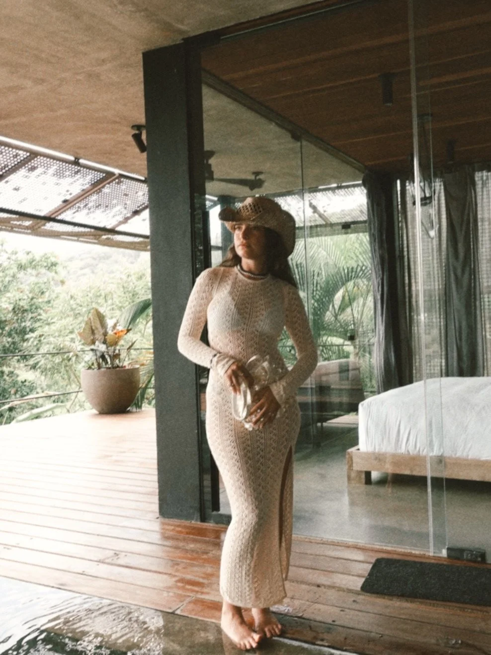Woman in a beige crochet dress and wide-brimmed hat holding a glass of water, standing on a wooden floor near a glass wall with outdoor greenery visible.
