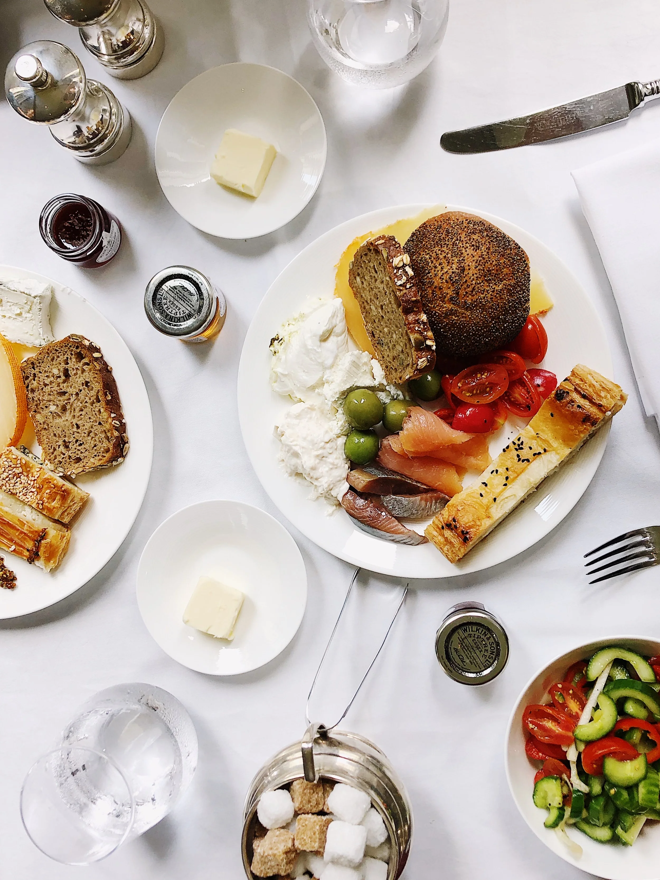 Table with assorted breakfast foods including bread, tomatoes, cucumbers, cheese, fish, and salad. There are also butter, jam, and condiments, along with glasses of water and sugar cubes.