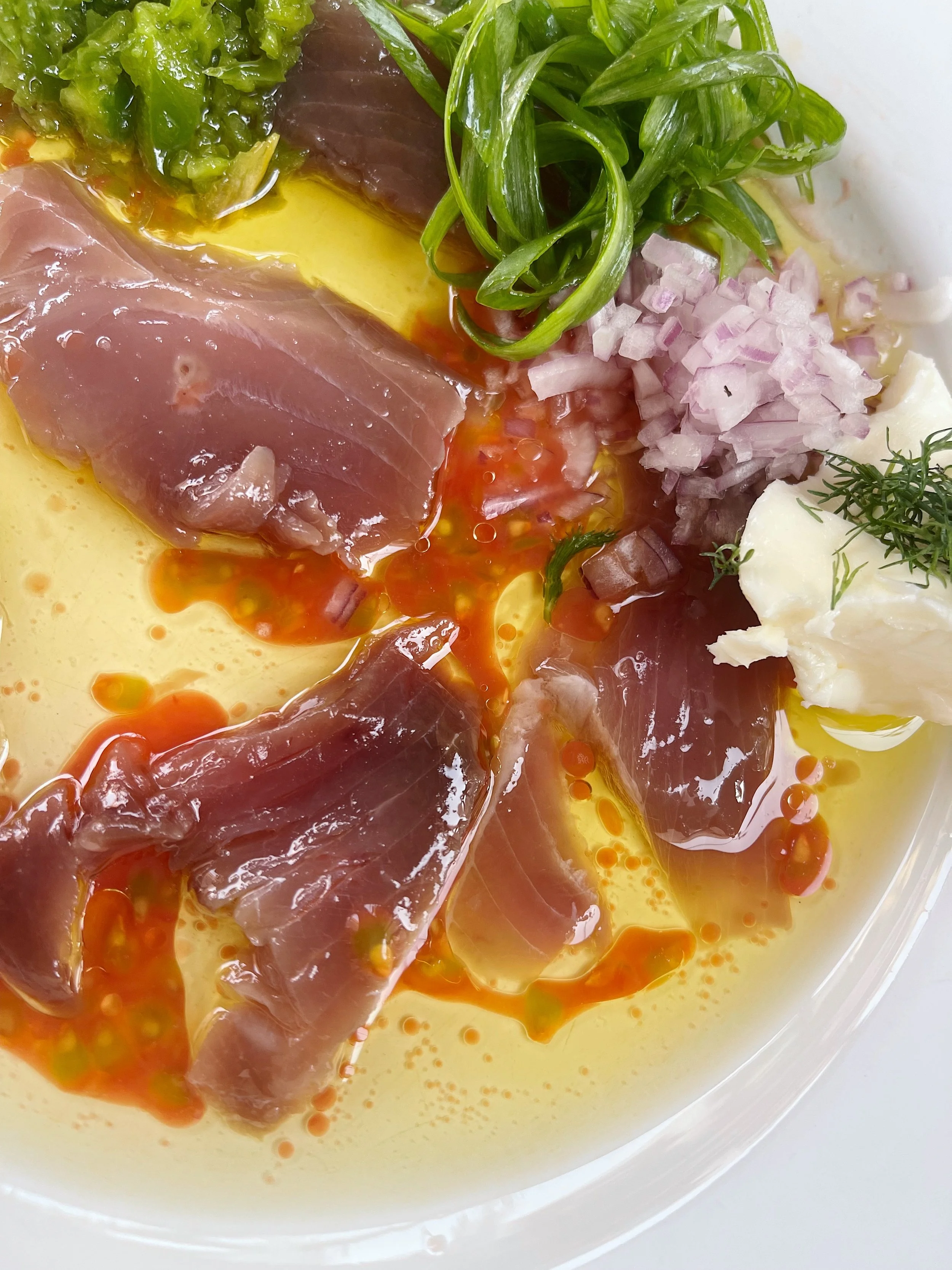Close-up of prepped ingredients for a dish, including raw fish fillets, chopped onions, leafy greens, chopped herbs, and a yellow sauce with red oil droplets.