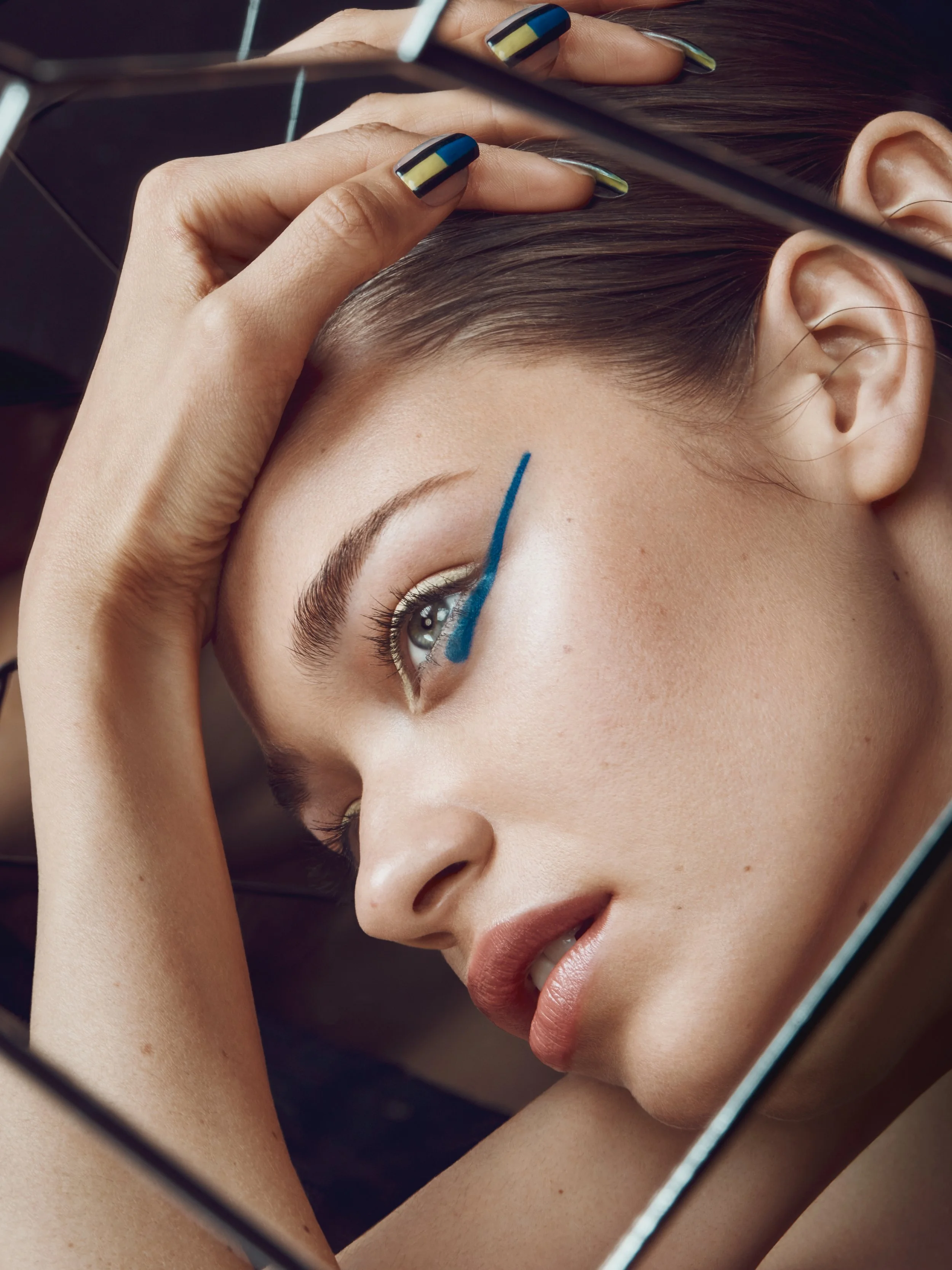 A woman with makeup and colored nails looking into a mirror, touching her head, with blue makeup eyeliner.
