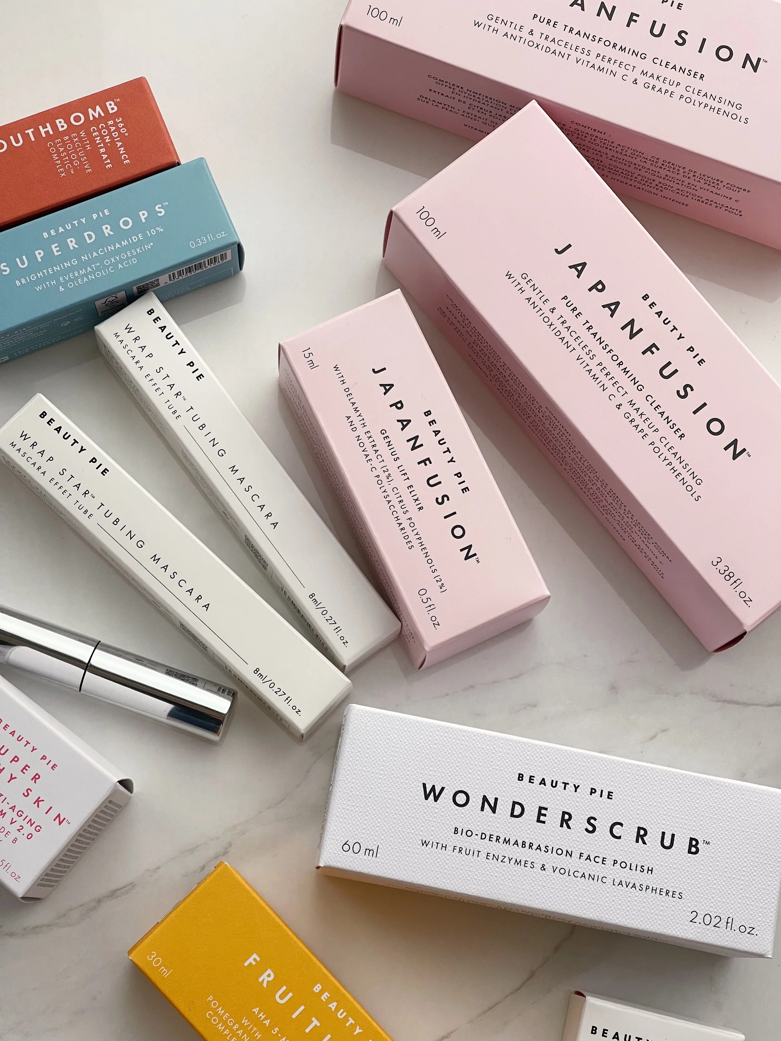 Various skincare and makeup products on a white marble surface, including boxes labeled 'Beauty Pie Superdrops', 'Japanfusion', 'Wonderscrub', and 'Fruit Vitamin C'. Items include mascara, face wash, and other beauty tools.