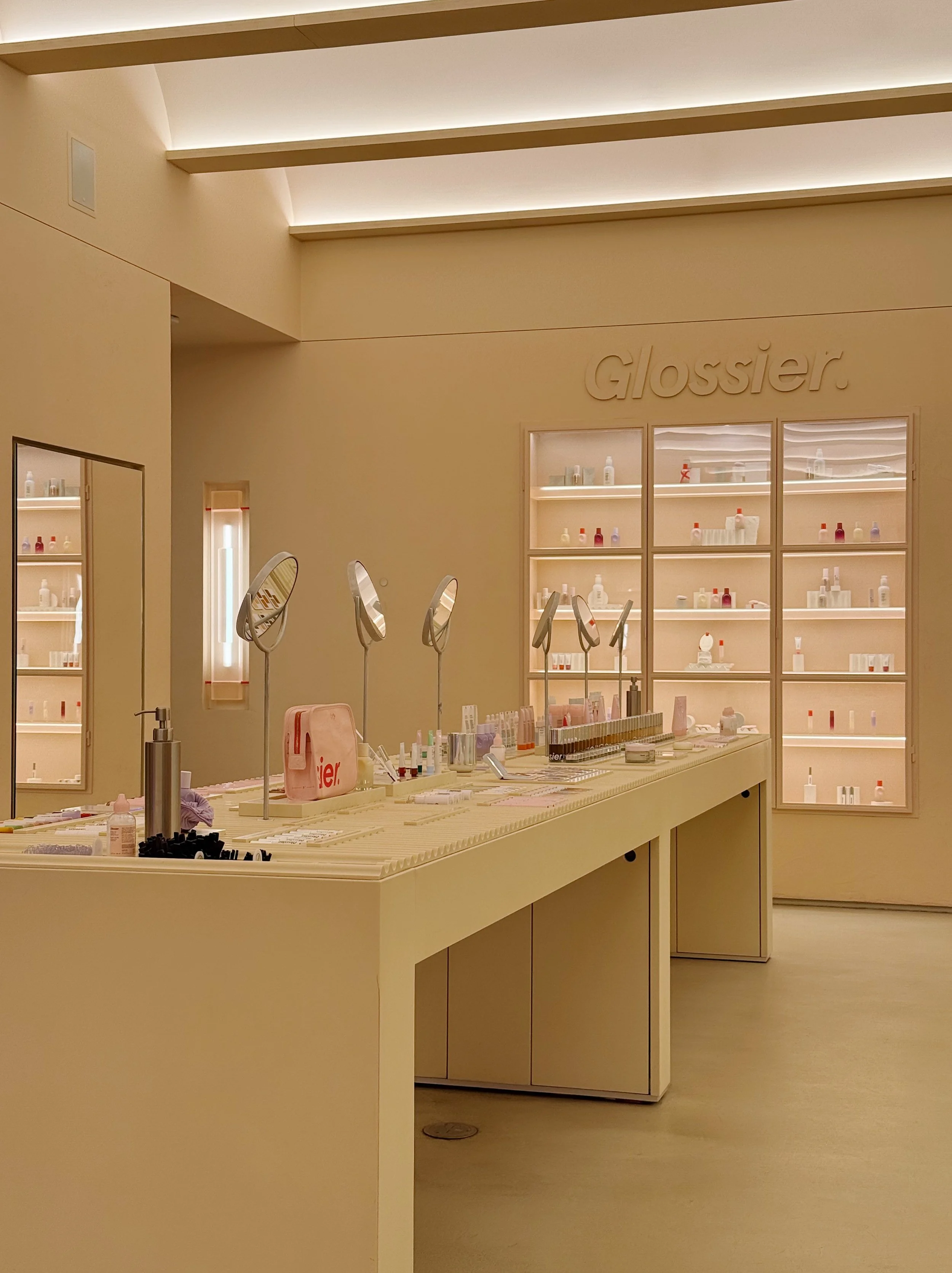 A minimalist cosmetics store display with shelves of skincare and beauty products, illuminated with soft lighting. The store's name, 'Glossier,' is embossed on the wall, emphasizing a modern, clean aesthetic.