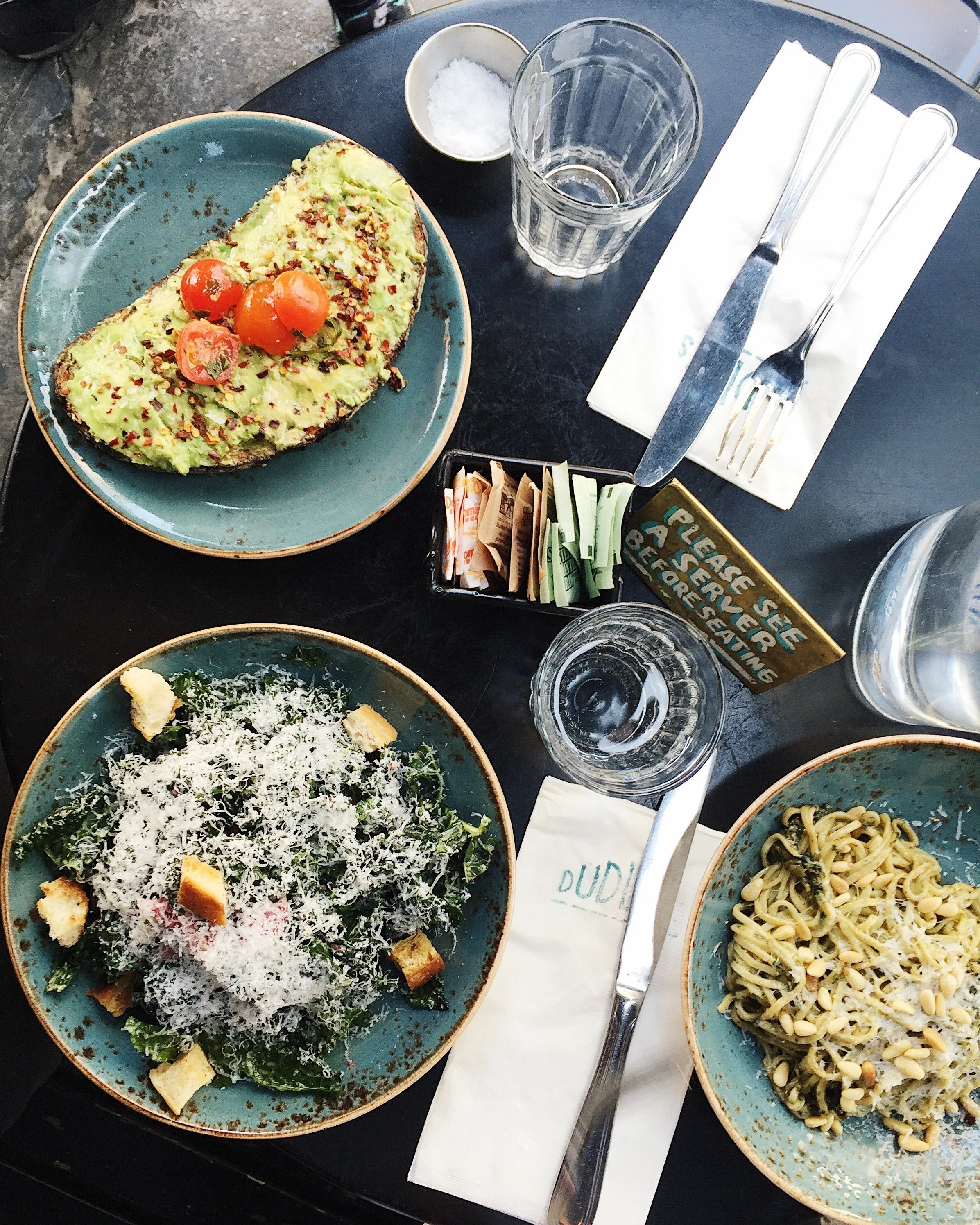 Table with three plates of food: toasted bread with avocado, cherry tomatoes, and chili flakes; Caesar salad with shredded cheese and croutons; pasta with pesto and pine nuts. Also has drinking glasses, salt, sugar packets, and utensils.