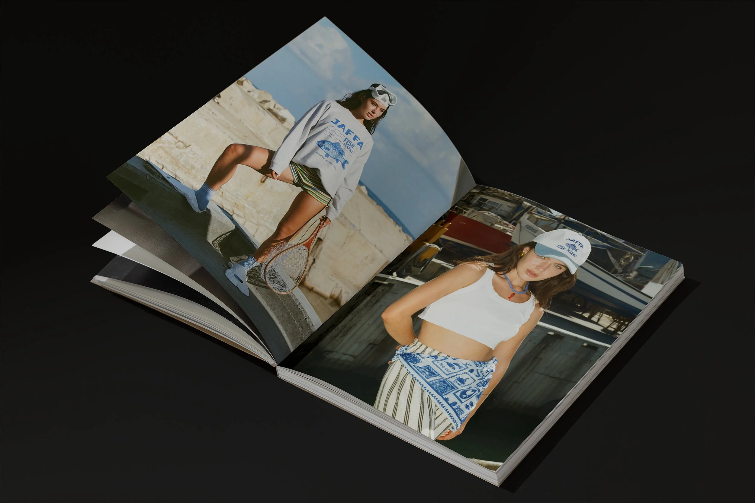 Open photo book with two images of women at a marina or dock, both in casual summer outfits, wearing caps with "JAFRA FISH MARKET" text, standing in front of boats and water.