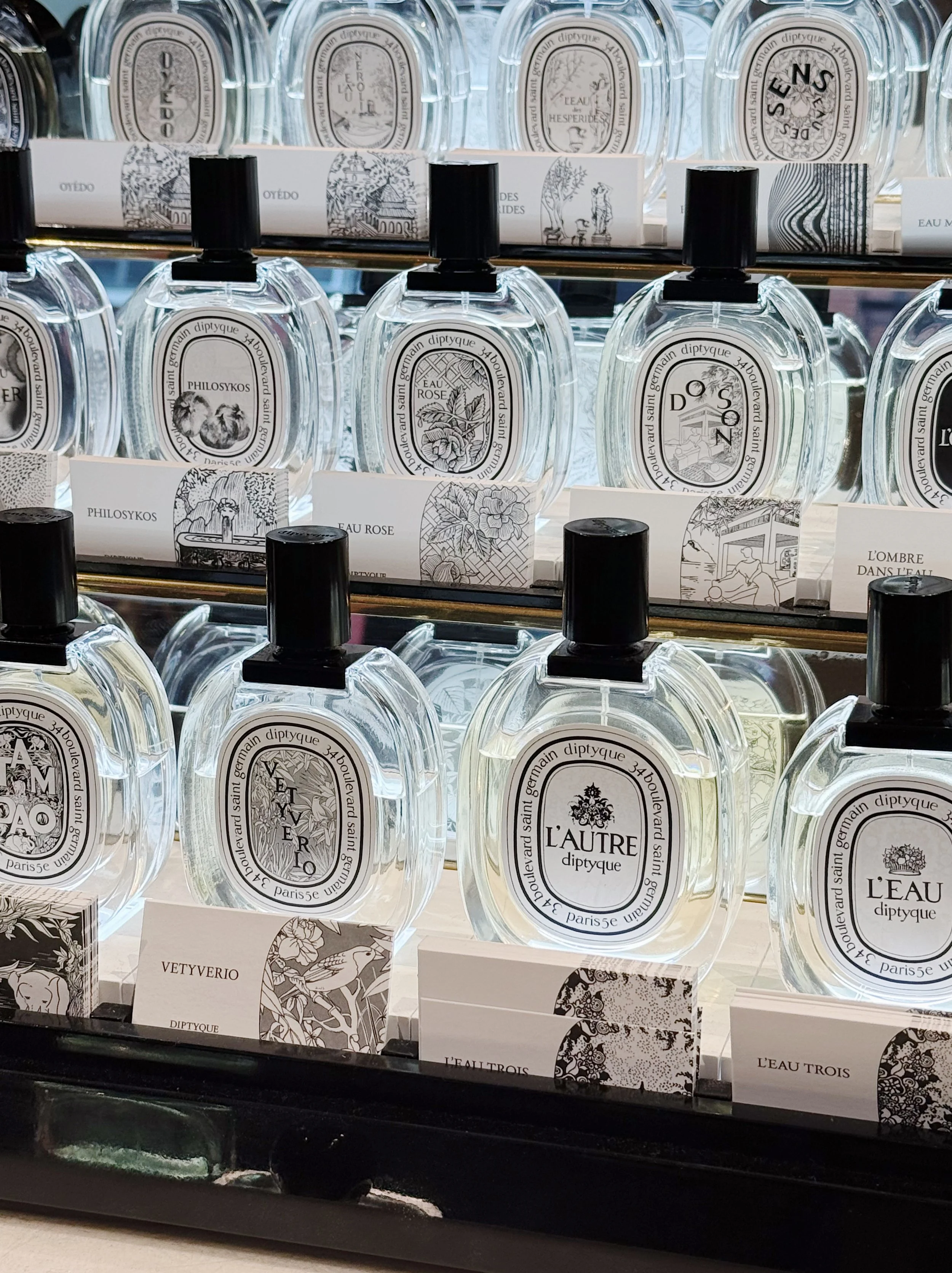 A display of various Diptyque perfume bottles on glass shelves, with labels featuring artistic designs and names like 'Vetyverio,' 'L'Autre,' and 'L'Eau Trois.'