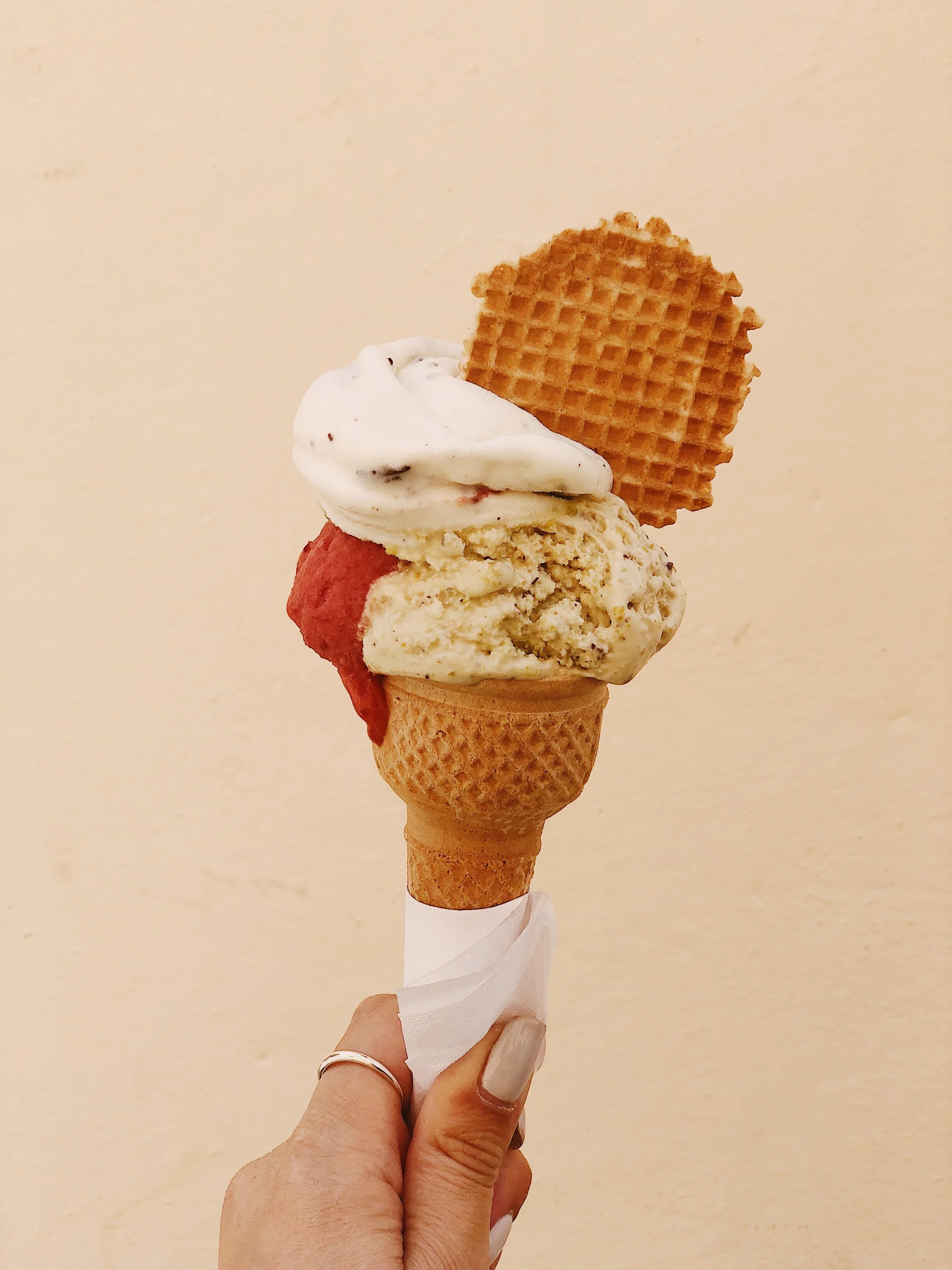 A hand holding an ice cream cone with three scoops topped with a waffle cookie, against a plain beige background.