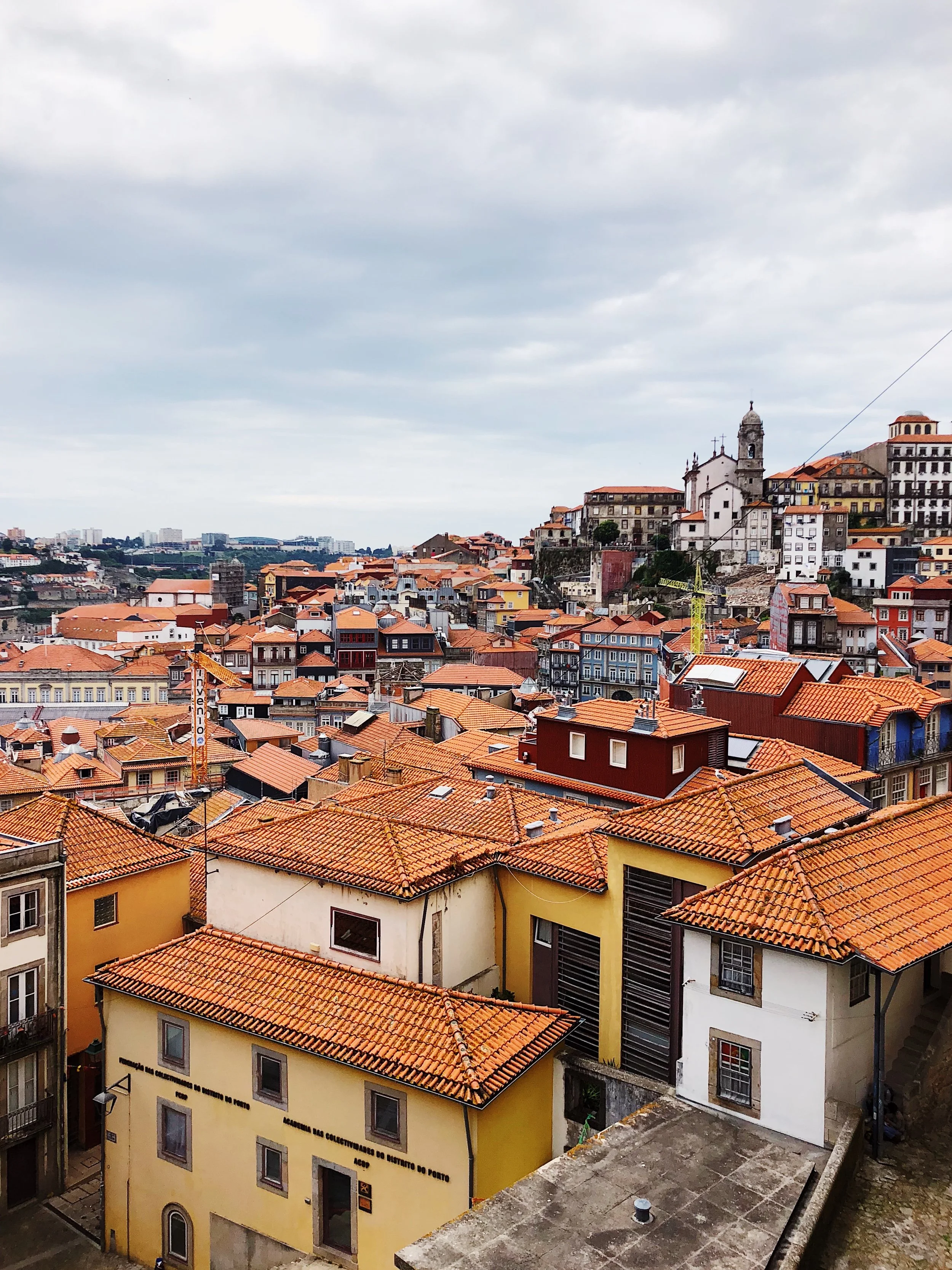View of a city with closely packed buildings with red-tiled roofs in an old European style, featuring a hillside with historic structures and a cloudy sky overhead.