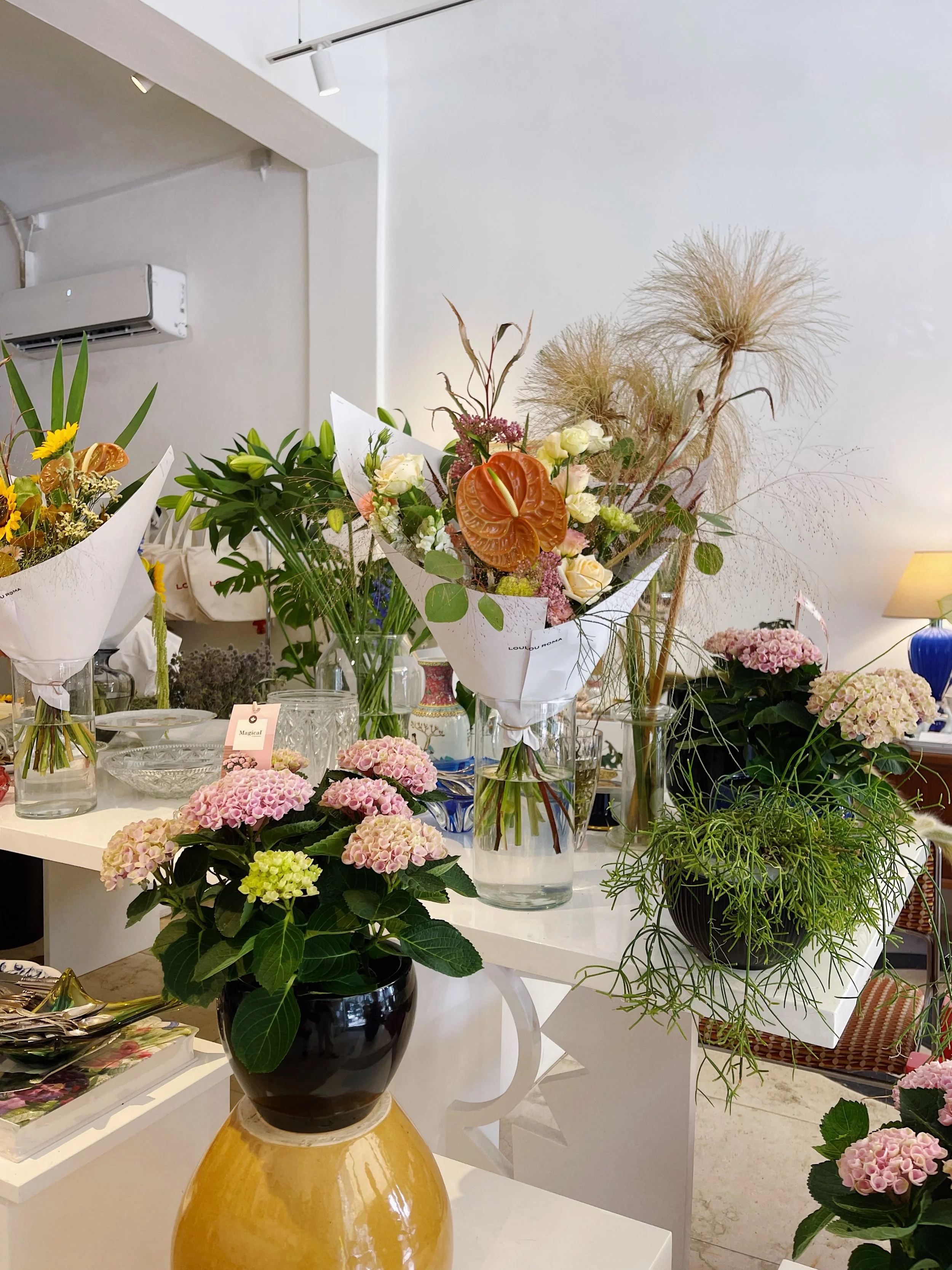 Arrangement of various flowers, including pink hydrangeas, white roses, and dried grasses, displayed on a white table in a bright room.