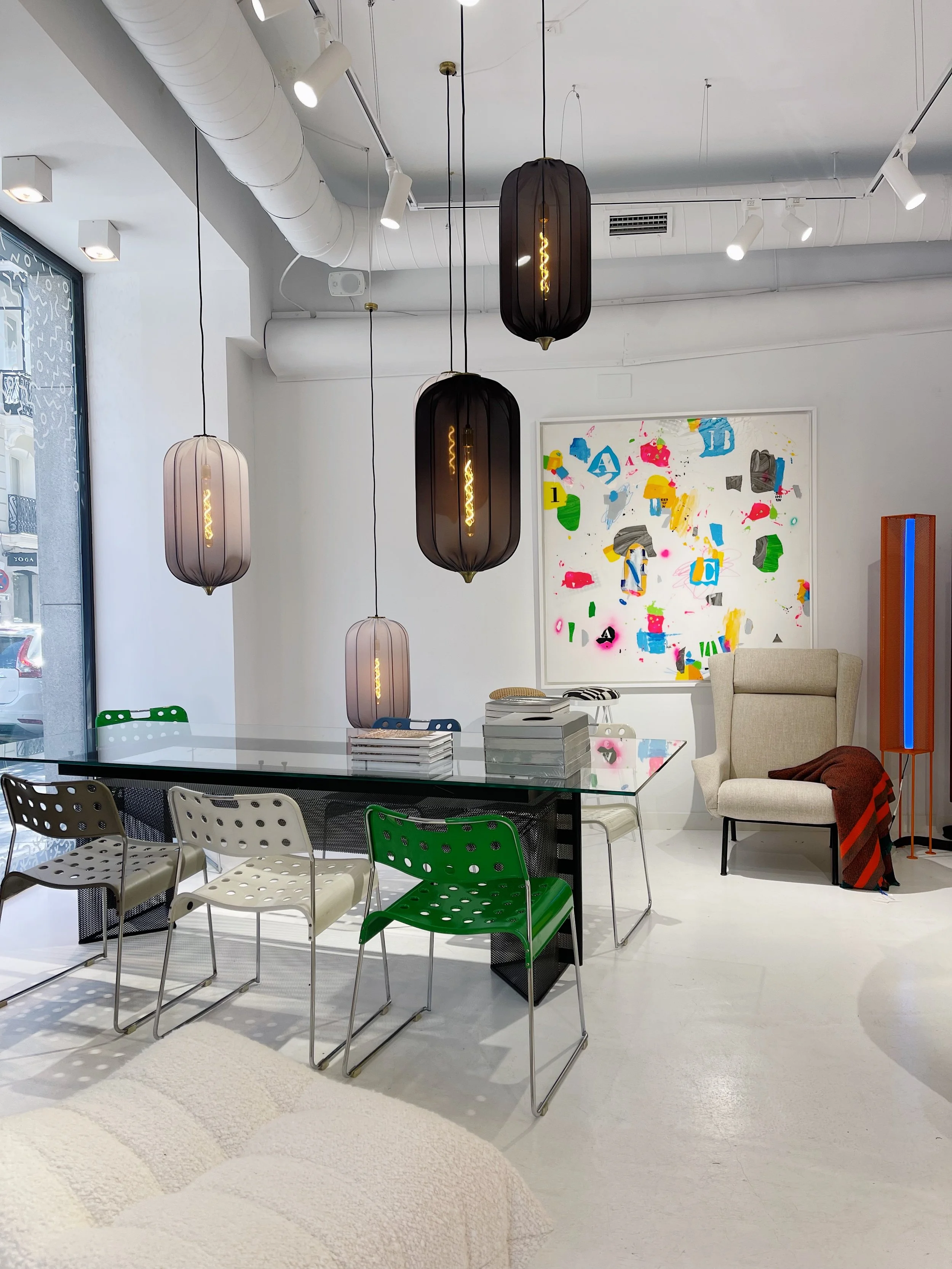 Modern, minimalistic office or meeting space with a glass table, white, beige, and green chairs, abstract artwork on the wall, hanging pendant lights, and a large window.