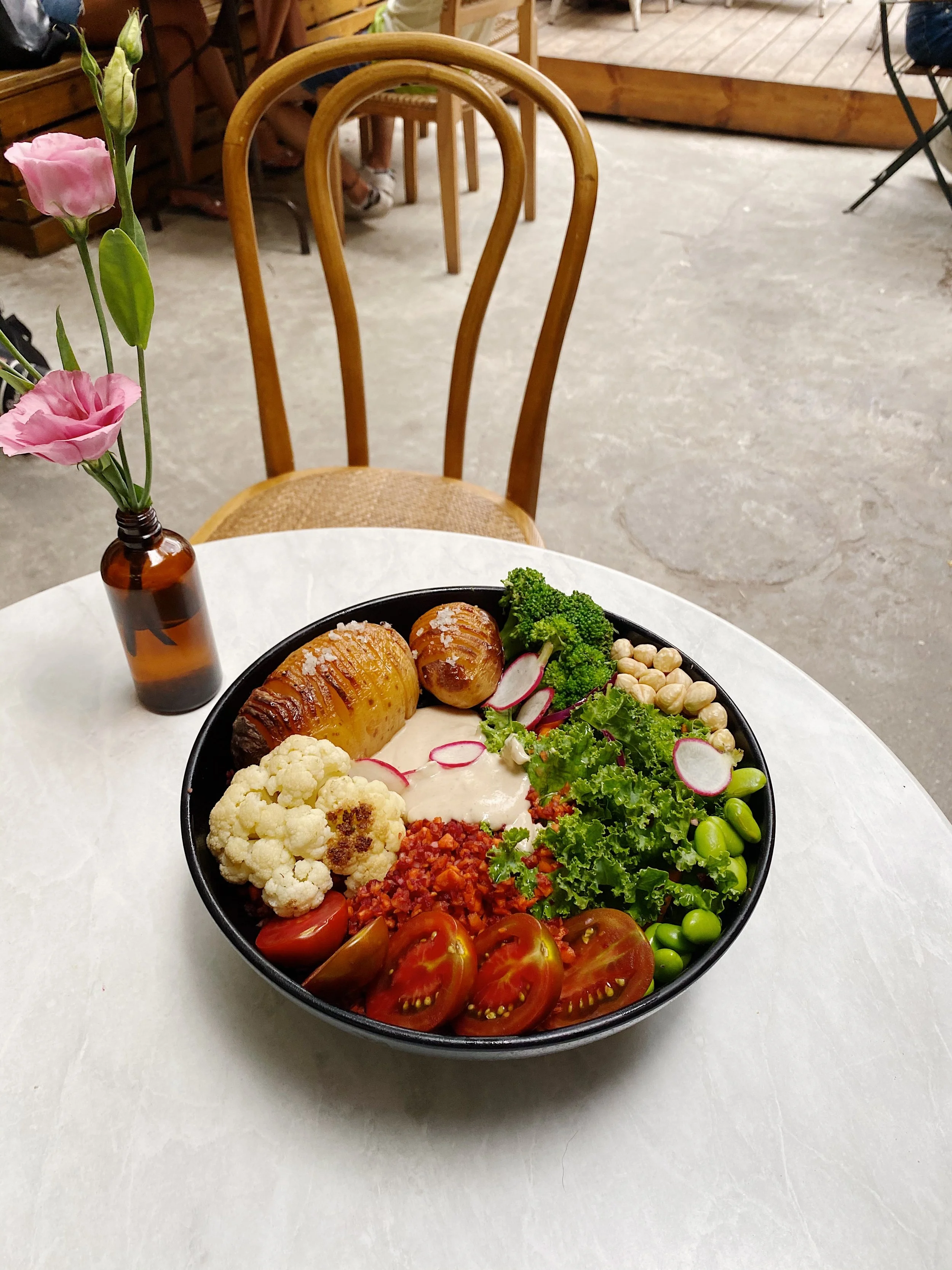 A bowl of mixed vegetables, including cherry tomatoes, cauliflower, broccoli, edamame, radishes, and hummus, on a white table with a small vase of pink flowers nearby.