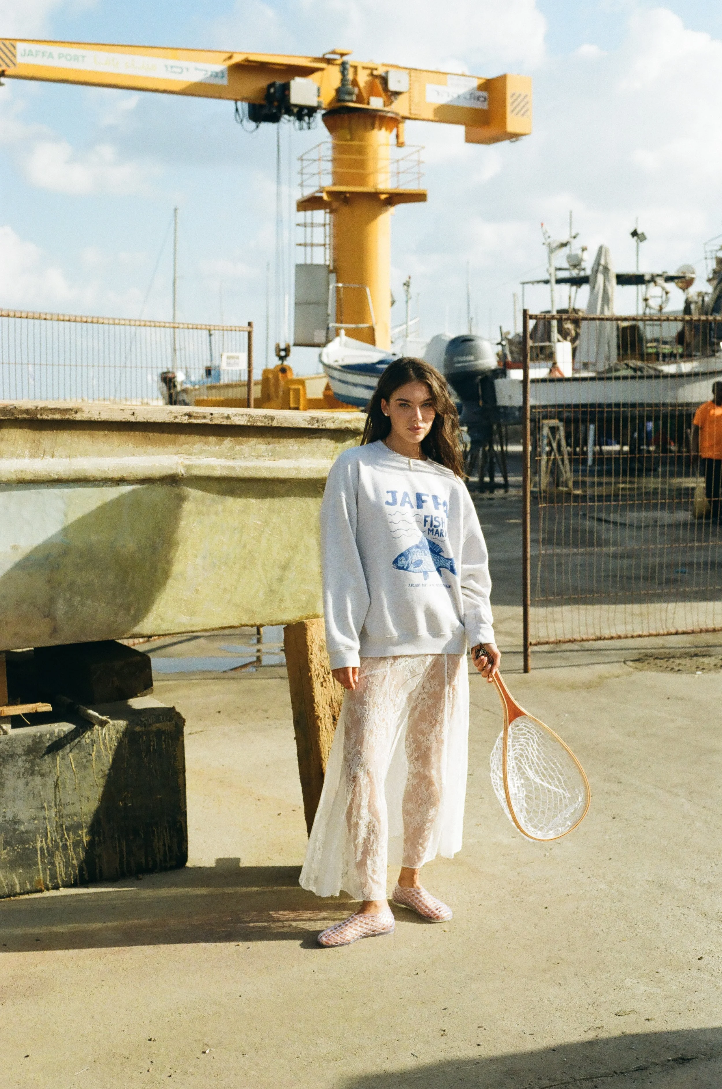A young woman standing near a boat dock, holding a tennis racket, dressed in a casual white sweatshirt and lace skirt, with boats and a yellow crane in the background.
