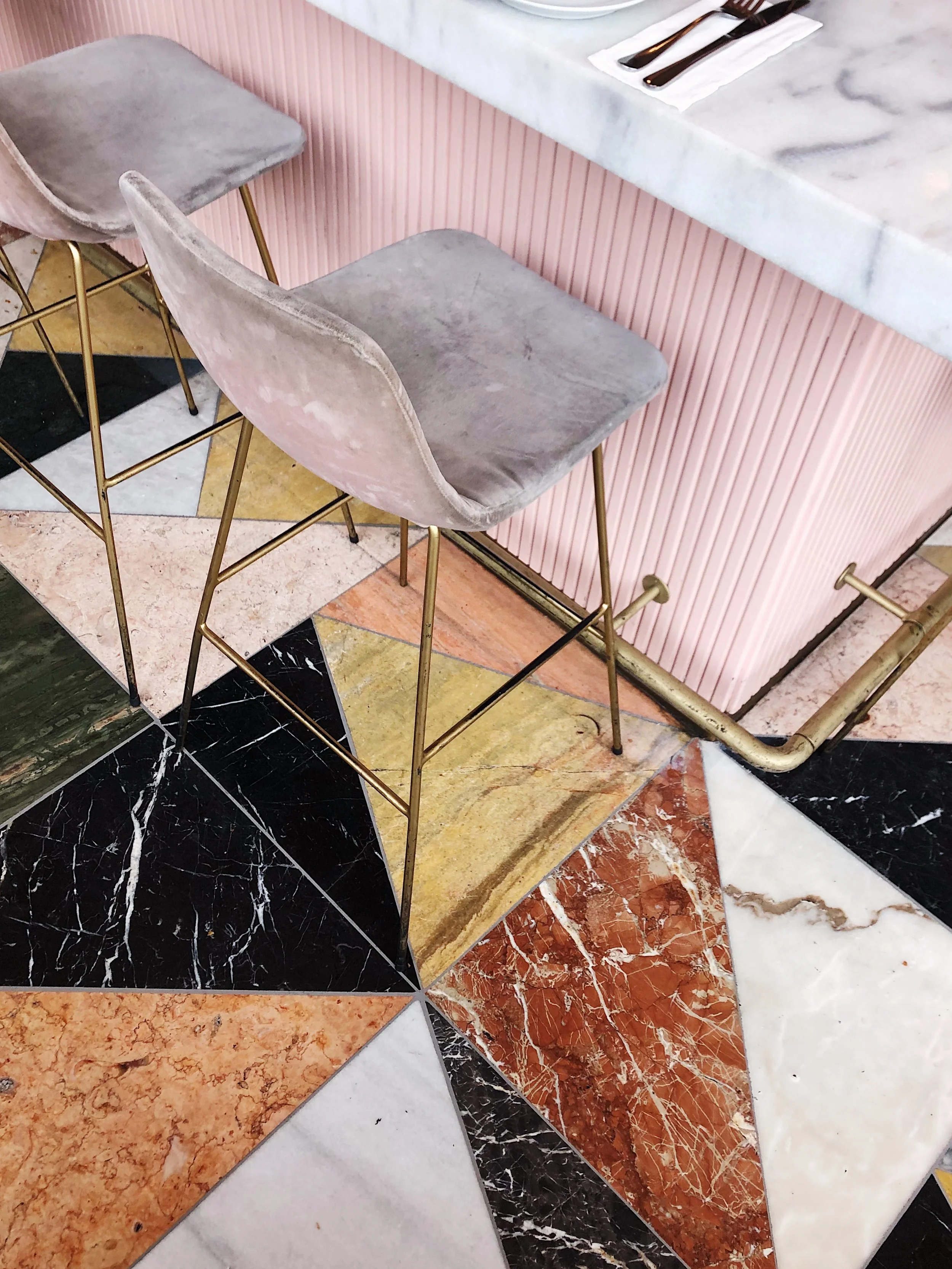 Gray velvet bar stools with gold legs in front of a pink textured bar counter, with a colorful patterned marble floor.