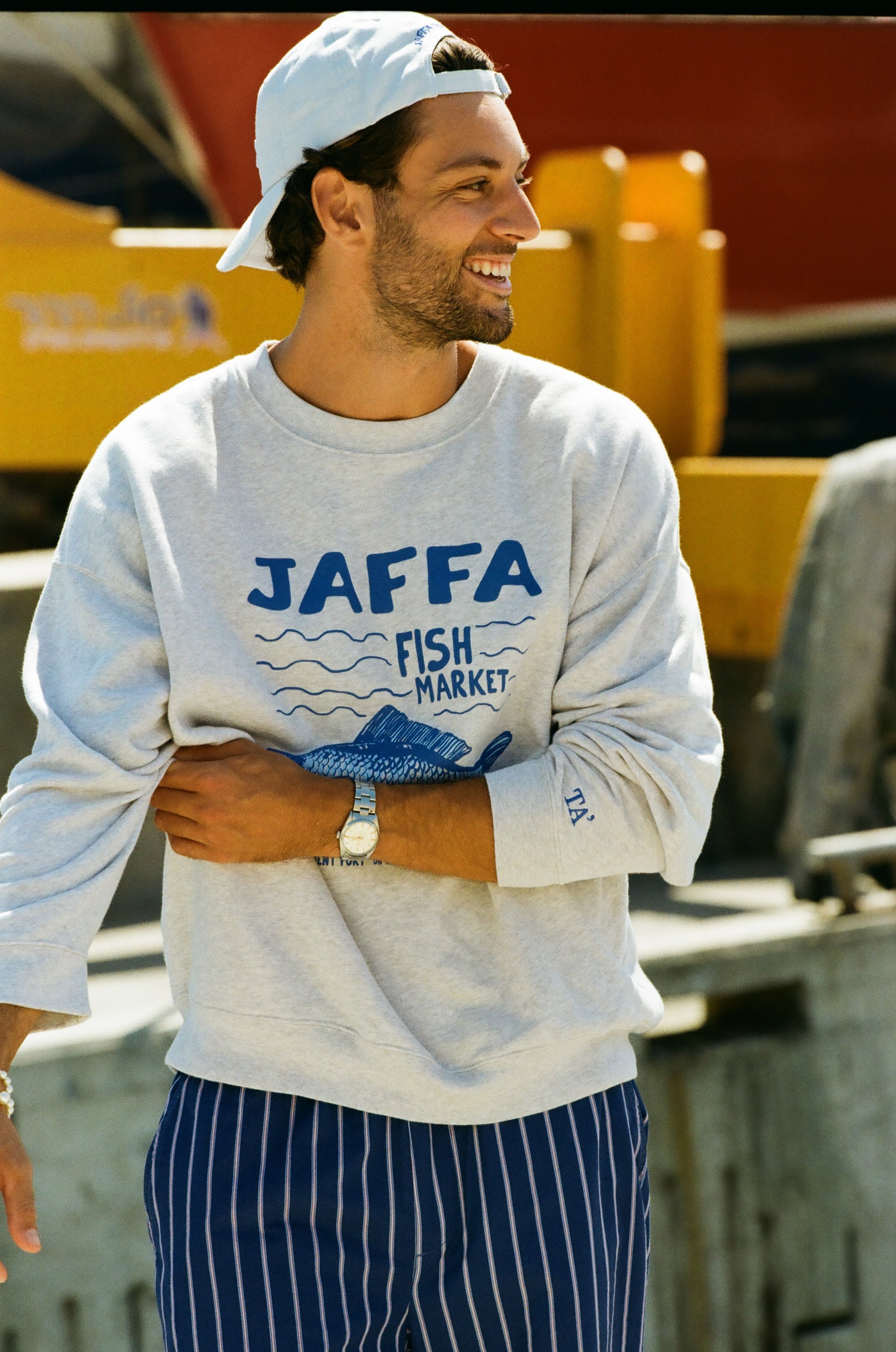 A man wearing a gray sweatshirt with blue text and graphics that say "Jaffa Fish Market," a white baseball cap worn backwards, a watch, and striped pants, standing outdoors near a fish market.