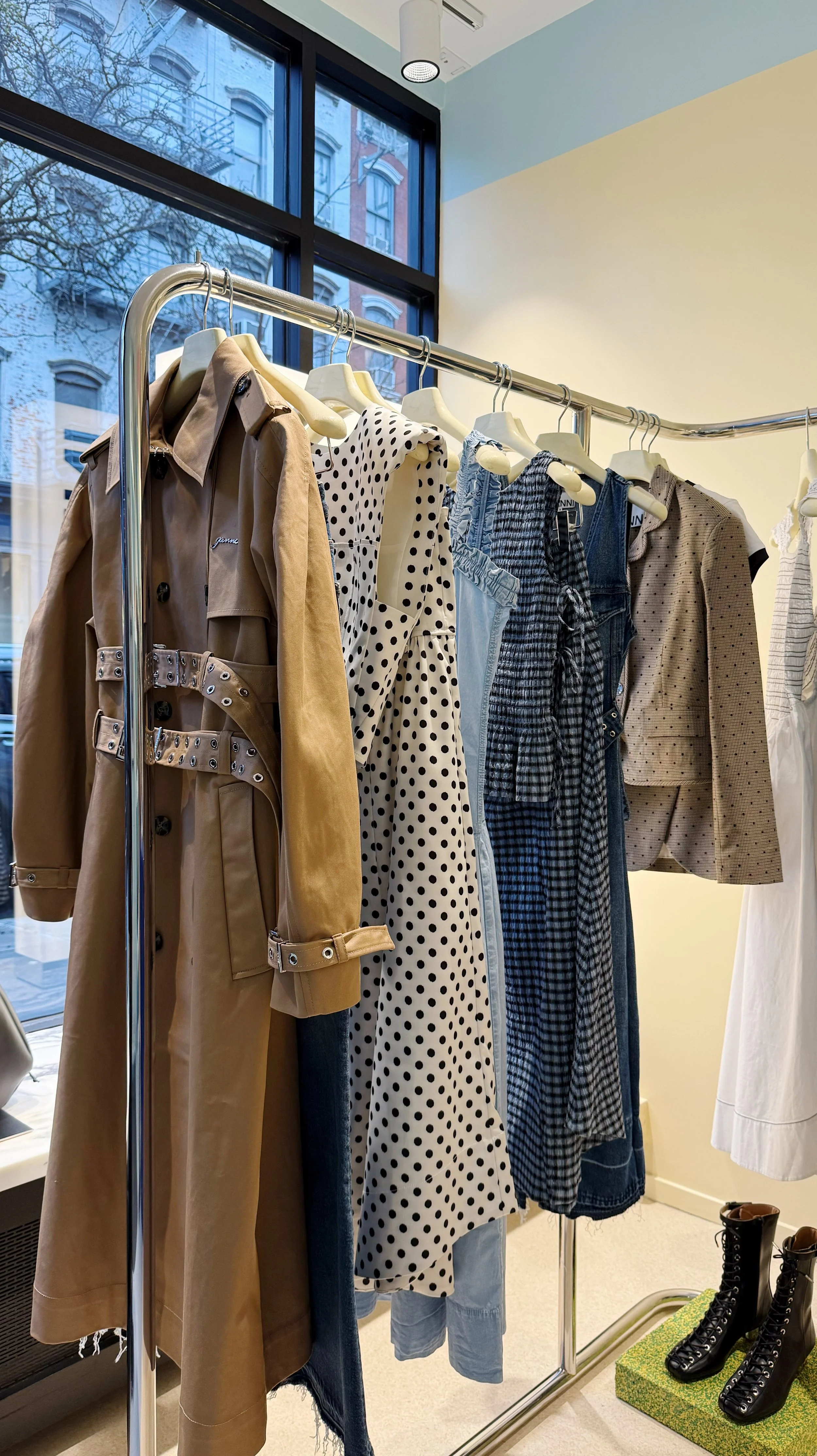 Clothing rack displaying various women's dresses, a coat, and a blazer in a store with a large window showing buildings outside.