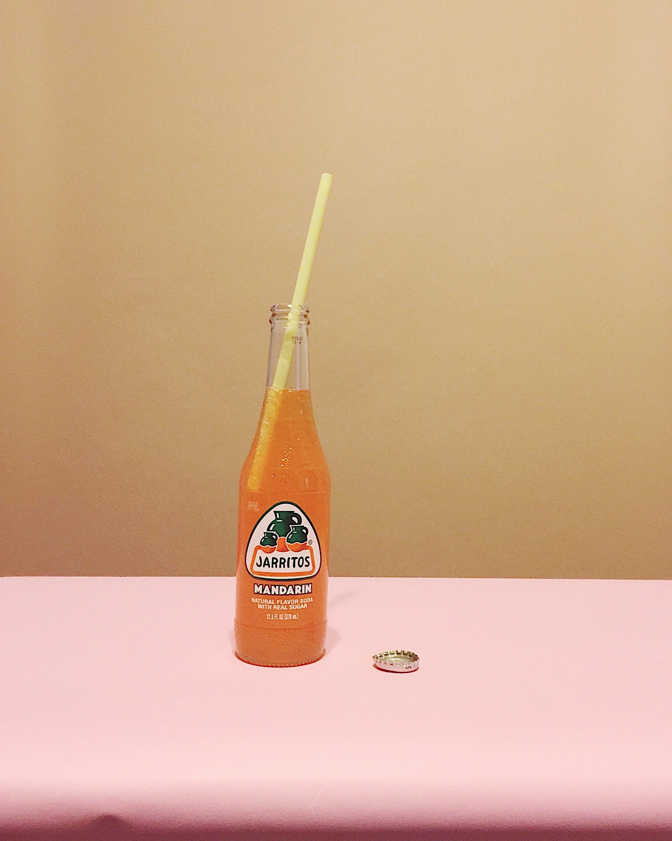 Bottle of Jarritos mandarin soda with a straw in it on a pink surface against a beige background, with a bottle cap on the surface.