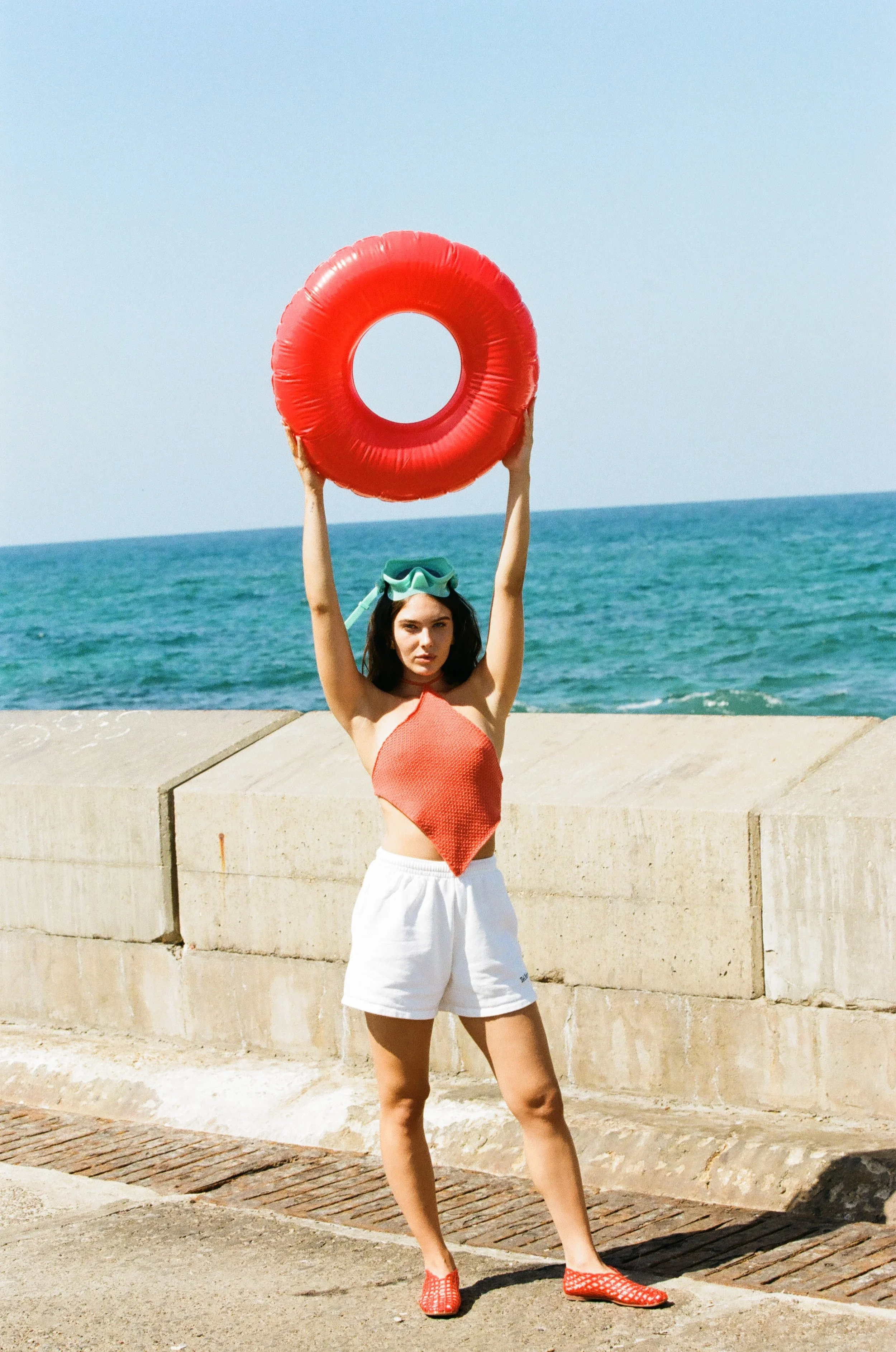 A woman standing on a pier at the beach, holding a red flotation ring above her head, wearing a red top, white shorts, and red shoes, with a green swim mask on her head, and the ocean in the background.