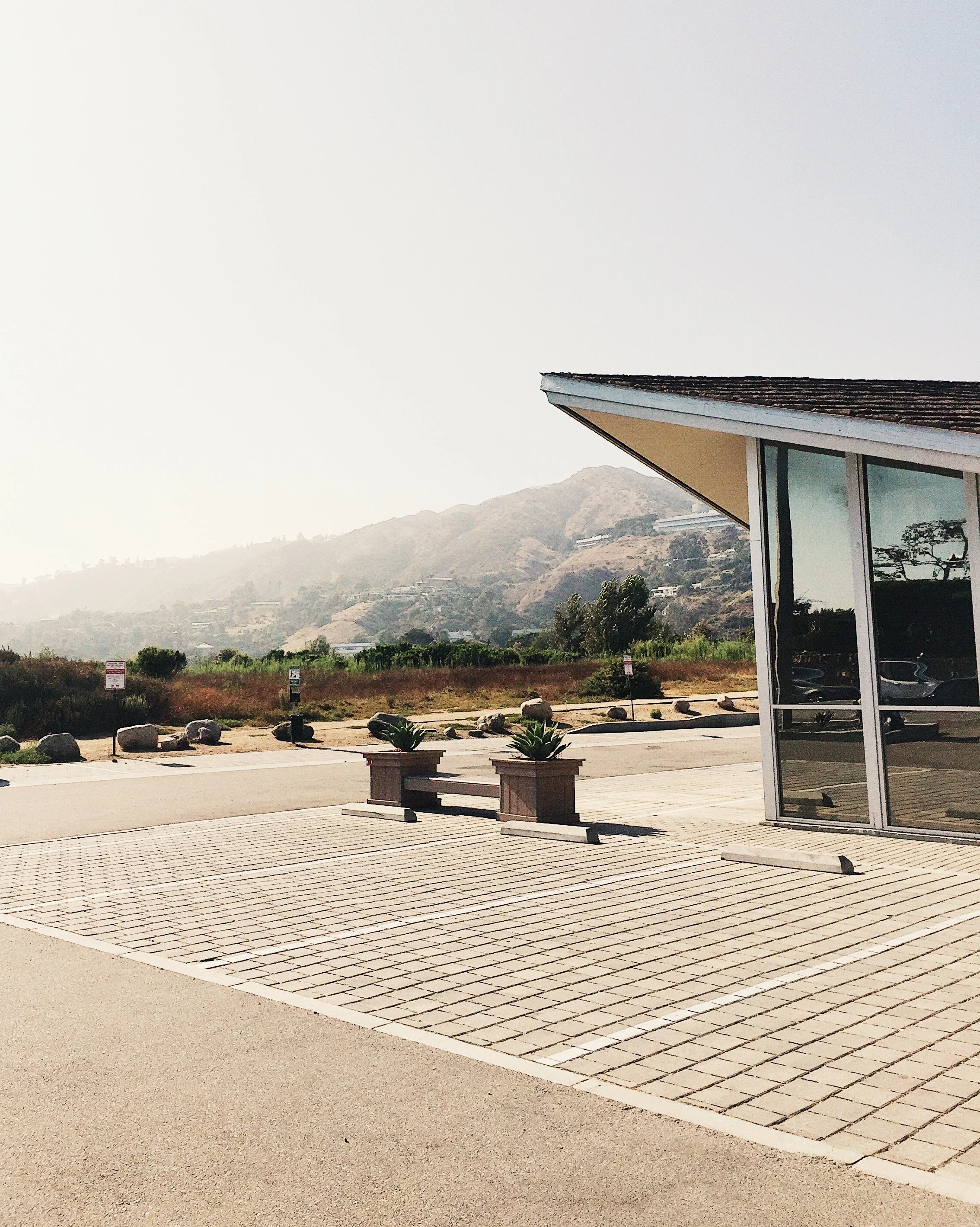 Empty parking lot outside a building with large windows, with potted plants and a mountain landscape in the background.