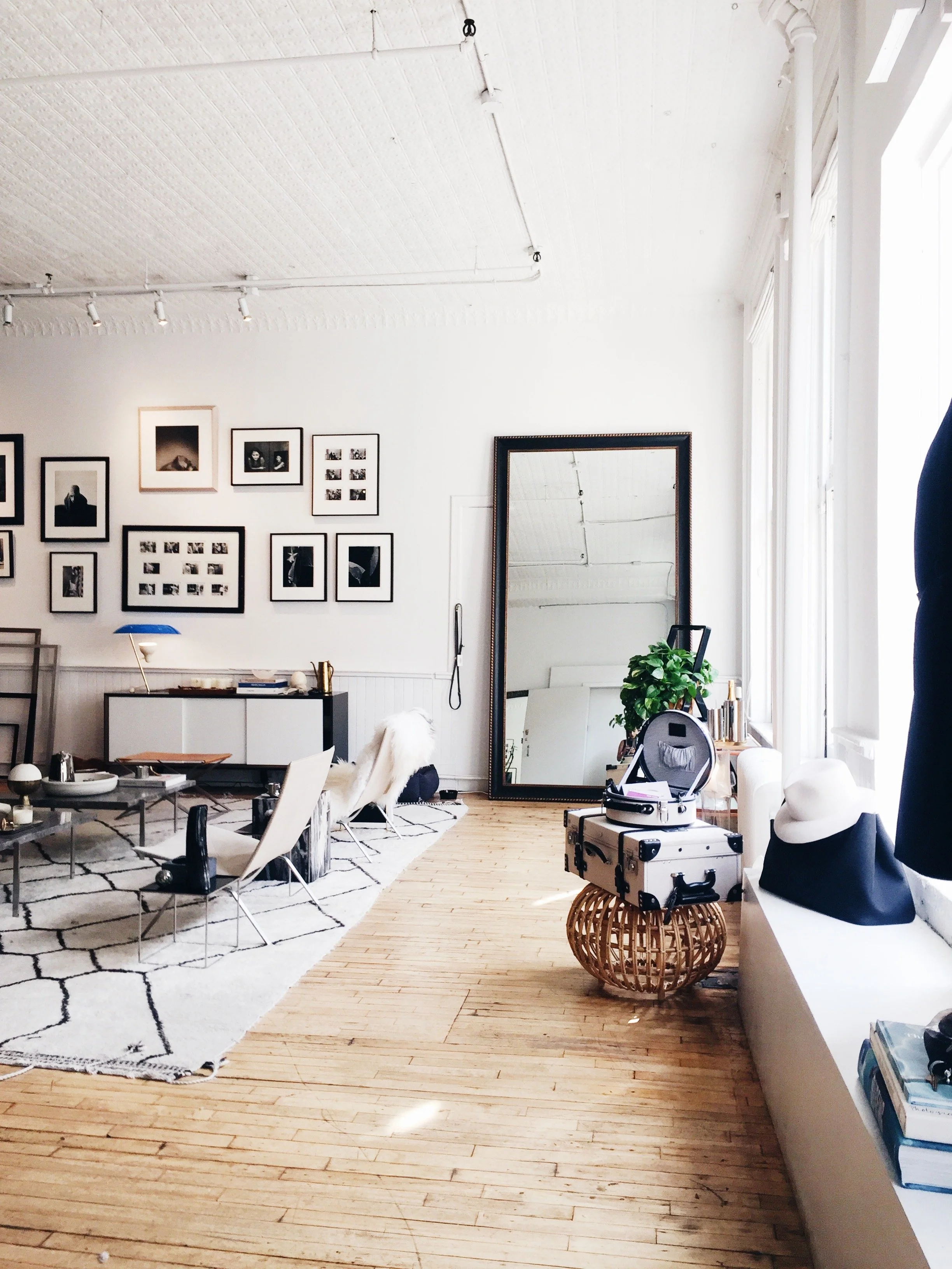 Bright living room with white walls, artwork, large mirror, wooden floor, and natural light. Decor includes plants, hats, and luggage.