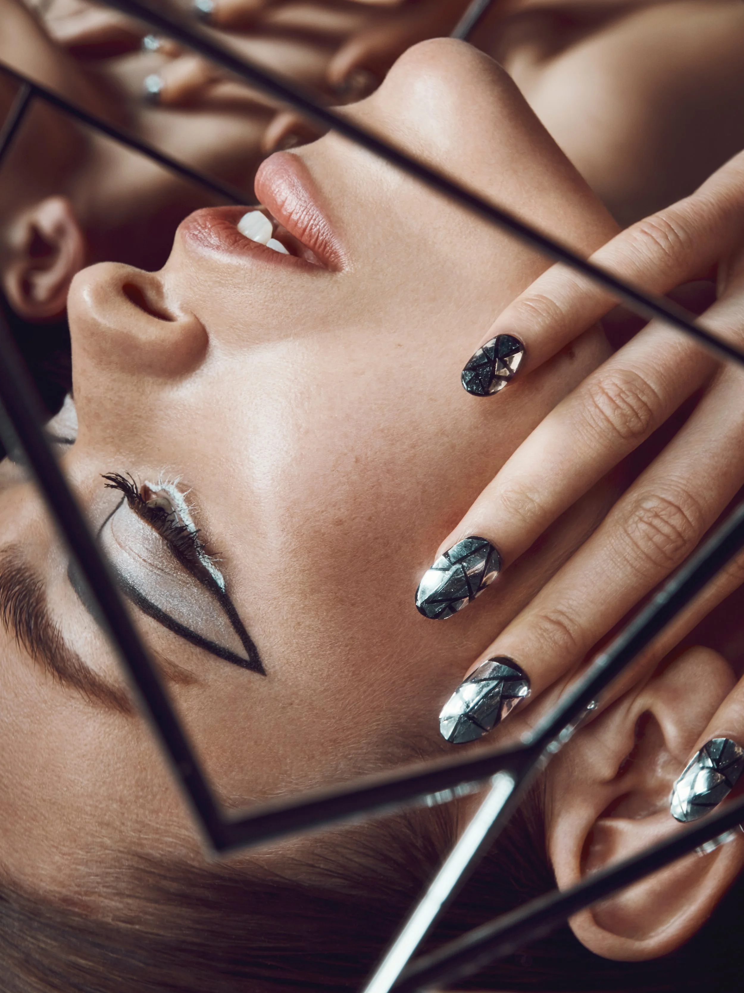 A person lying down with geometric makeup and matching nail art, surrounded by a reflective metallic frame.