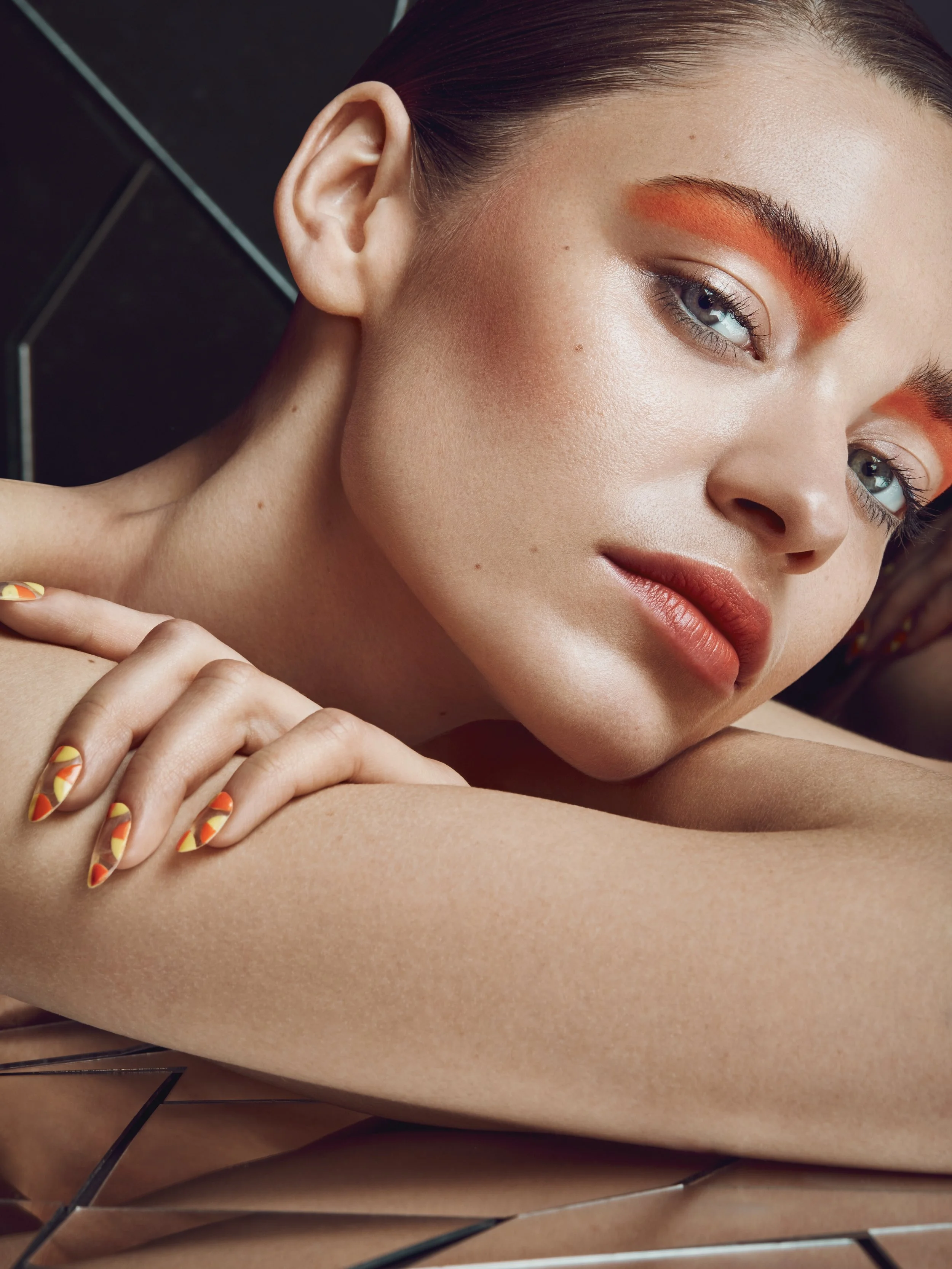 Close-up of a young woman with blue eyes, dark hair, bold orange makeup, and colorful manicure, resting her chin on her crossed arms against a dark tiled background.