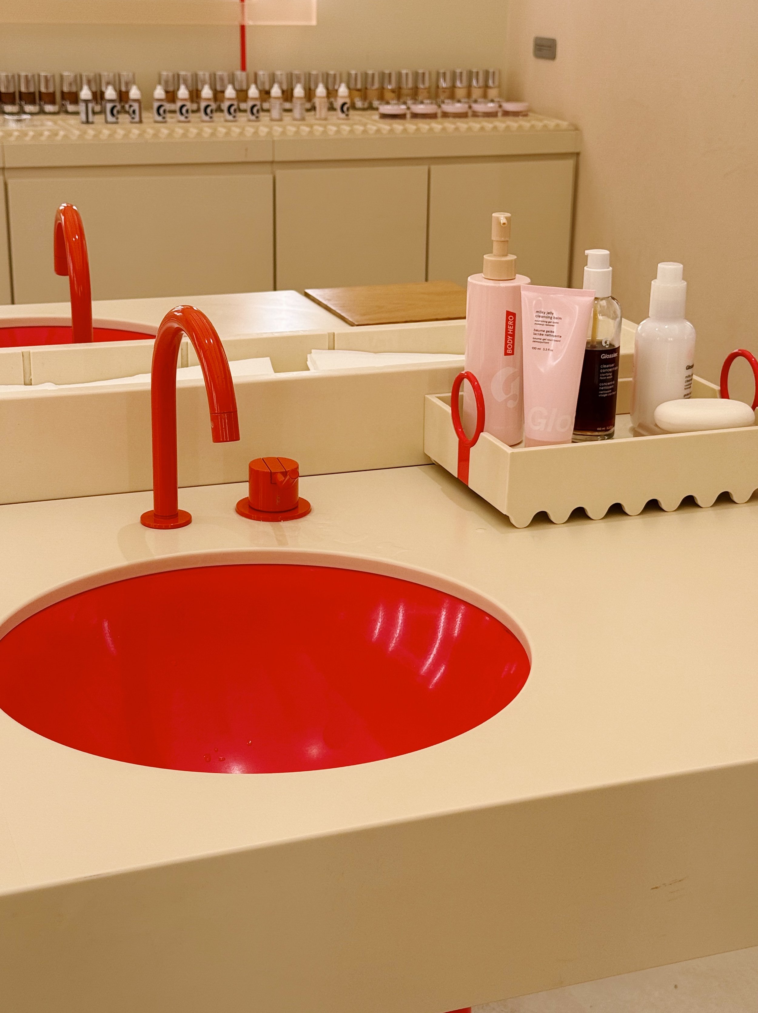 A bathroom or vanity area with a round red sink, red faucet, and a countertop with skincare and beauty products, including bottles and a bar soap. In the background, there are many small bottles and containers arranged on shelves.