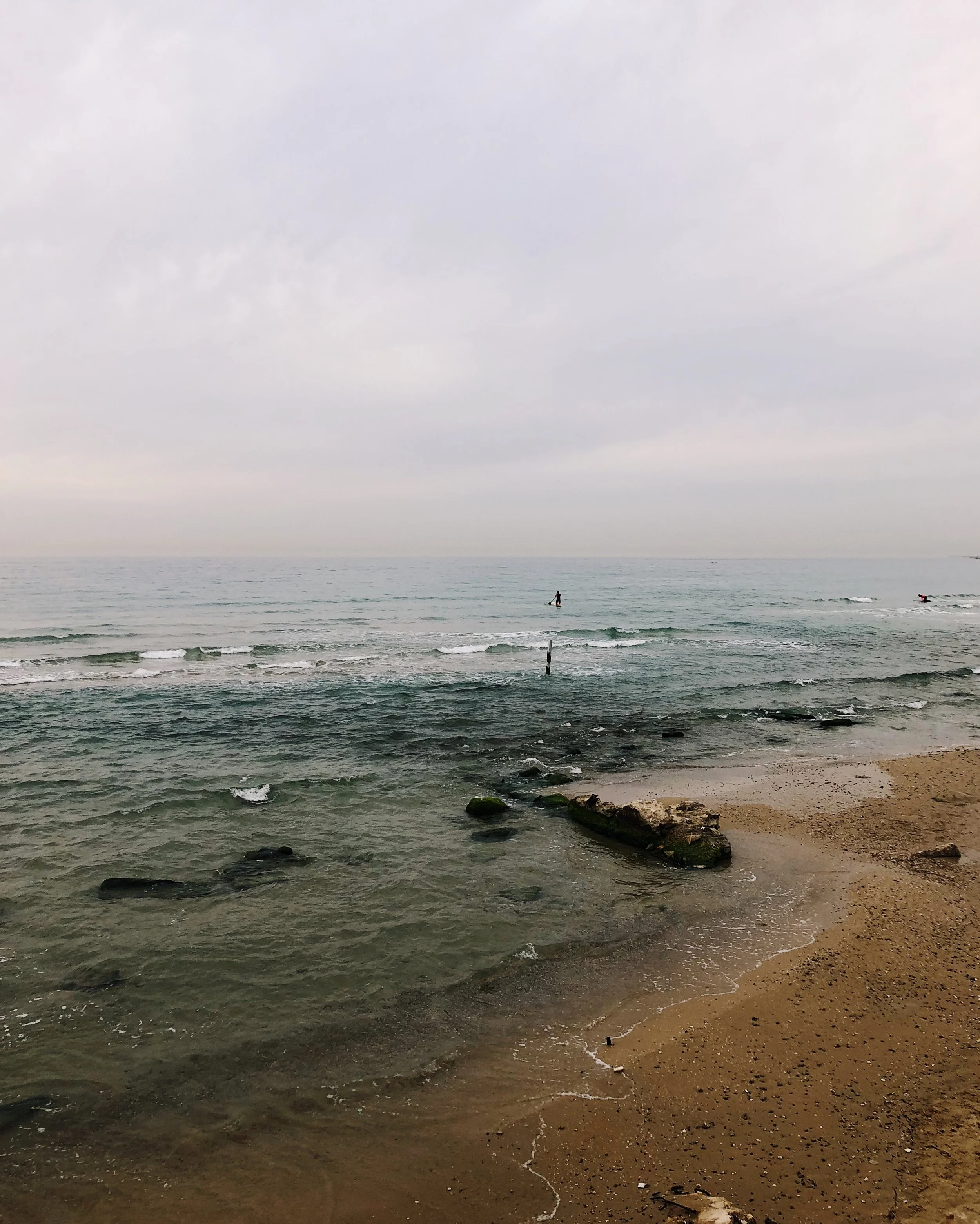 Beach with sand, rocks, and waves, with two surfers in the water and overcast sky.