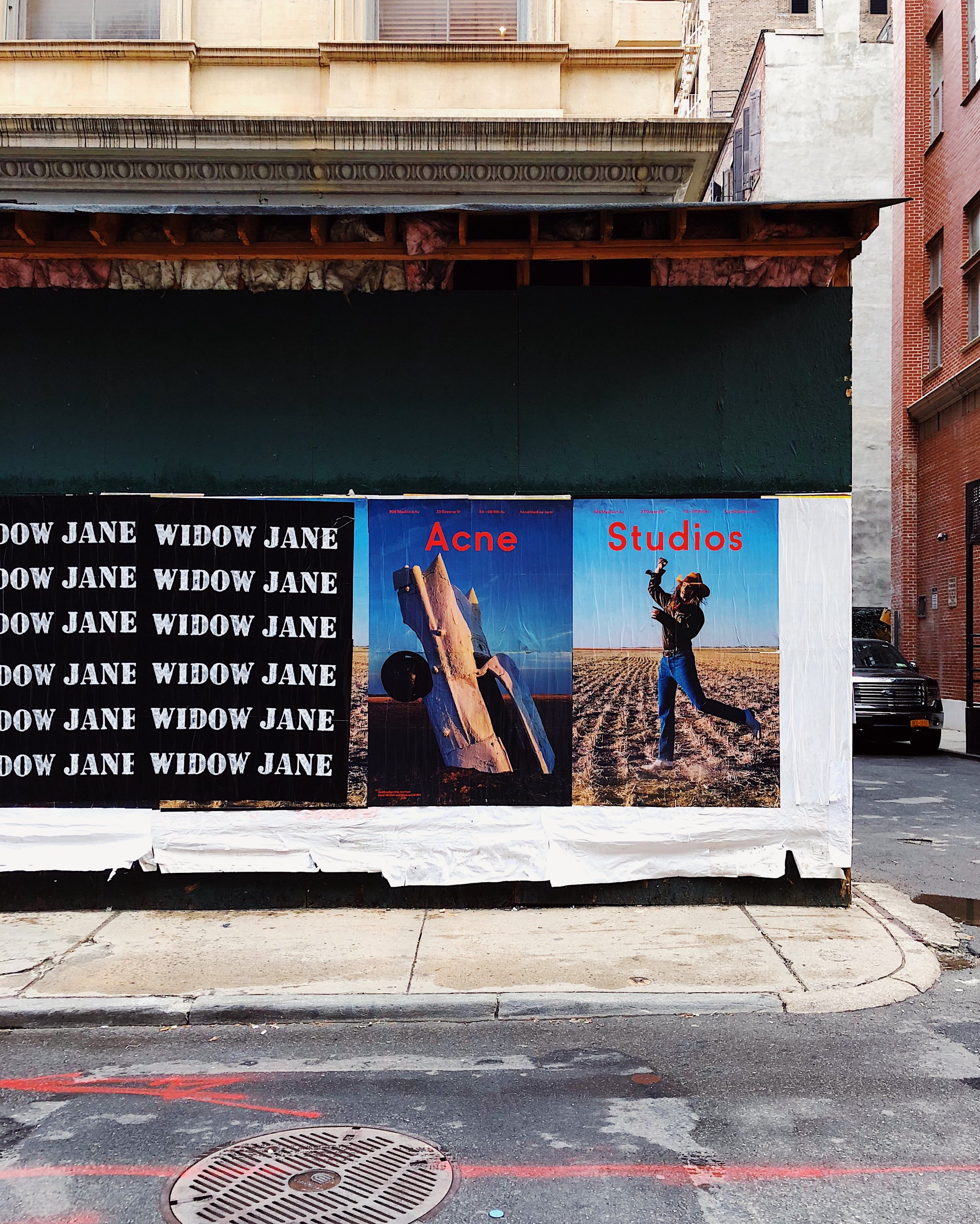 Street advertisement posters showing the words 'Widow Jane' repeated multiple times, with adjacent posters labeled 'Acne' and 'Studios'. The 'Acne' poster features a large piece of wood and a black circular object against a scenic background, and the