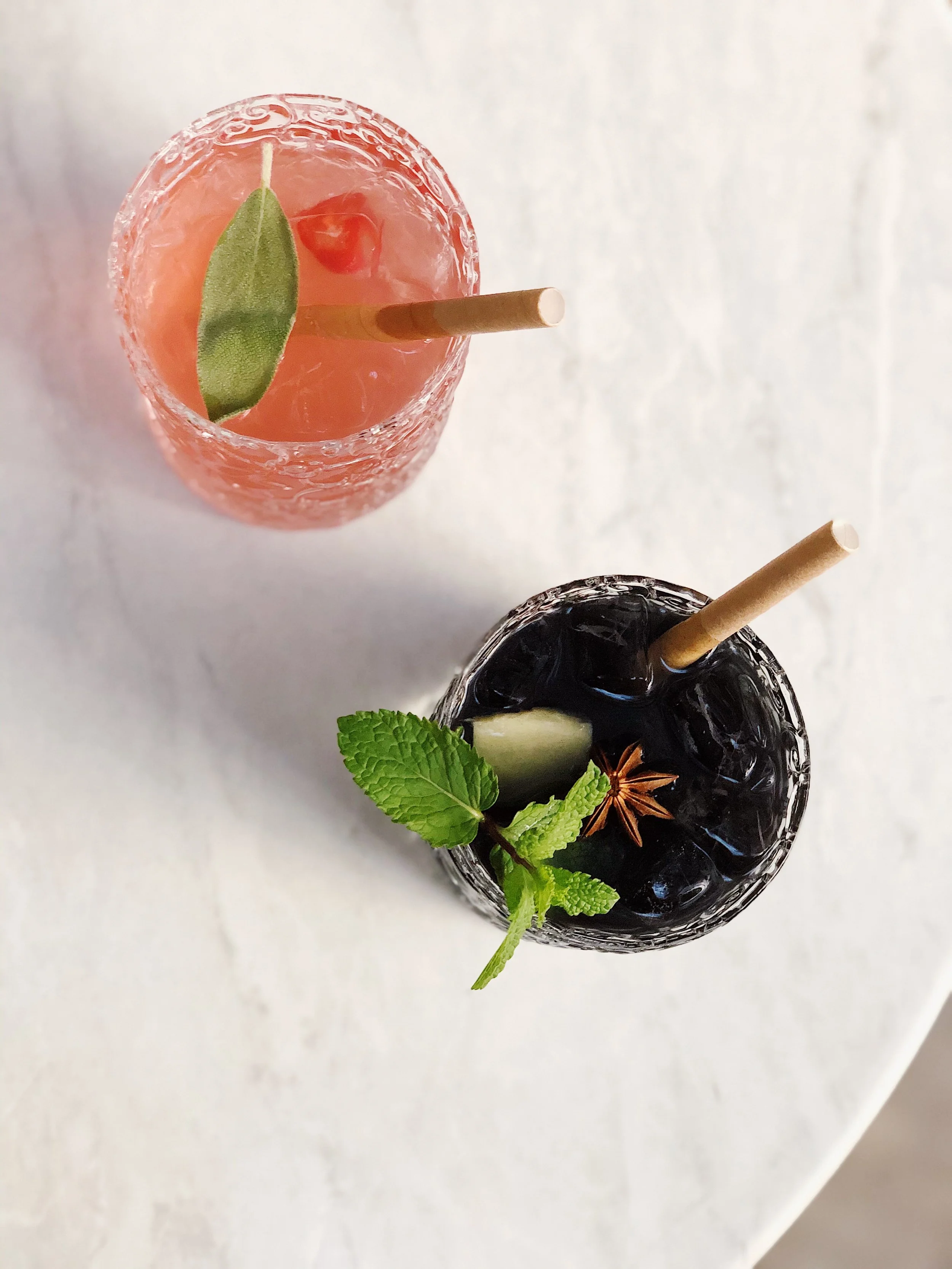 Two colorful cocktails on a white surface, one pink with a green leaf garnish and a straw, the other dark with mint, lemon, and star anise garnishes, each with a straw.