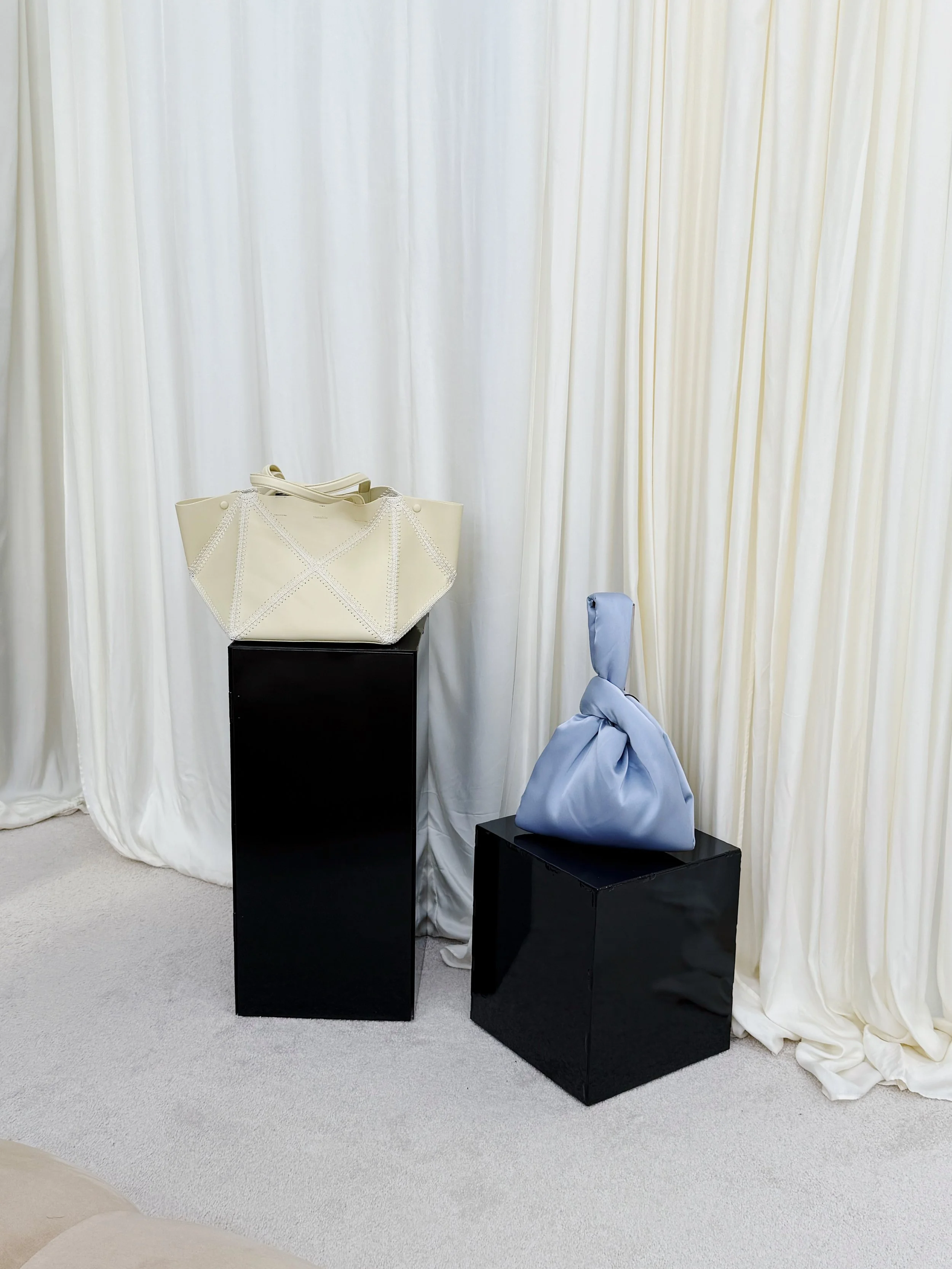 Two designer handbags, one cream and one light blue, displayed on black pedestal stands against a cream-colored curtain backdrop.