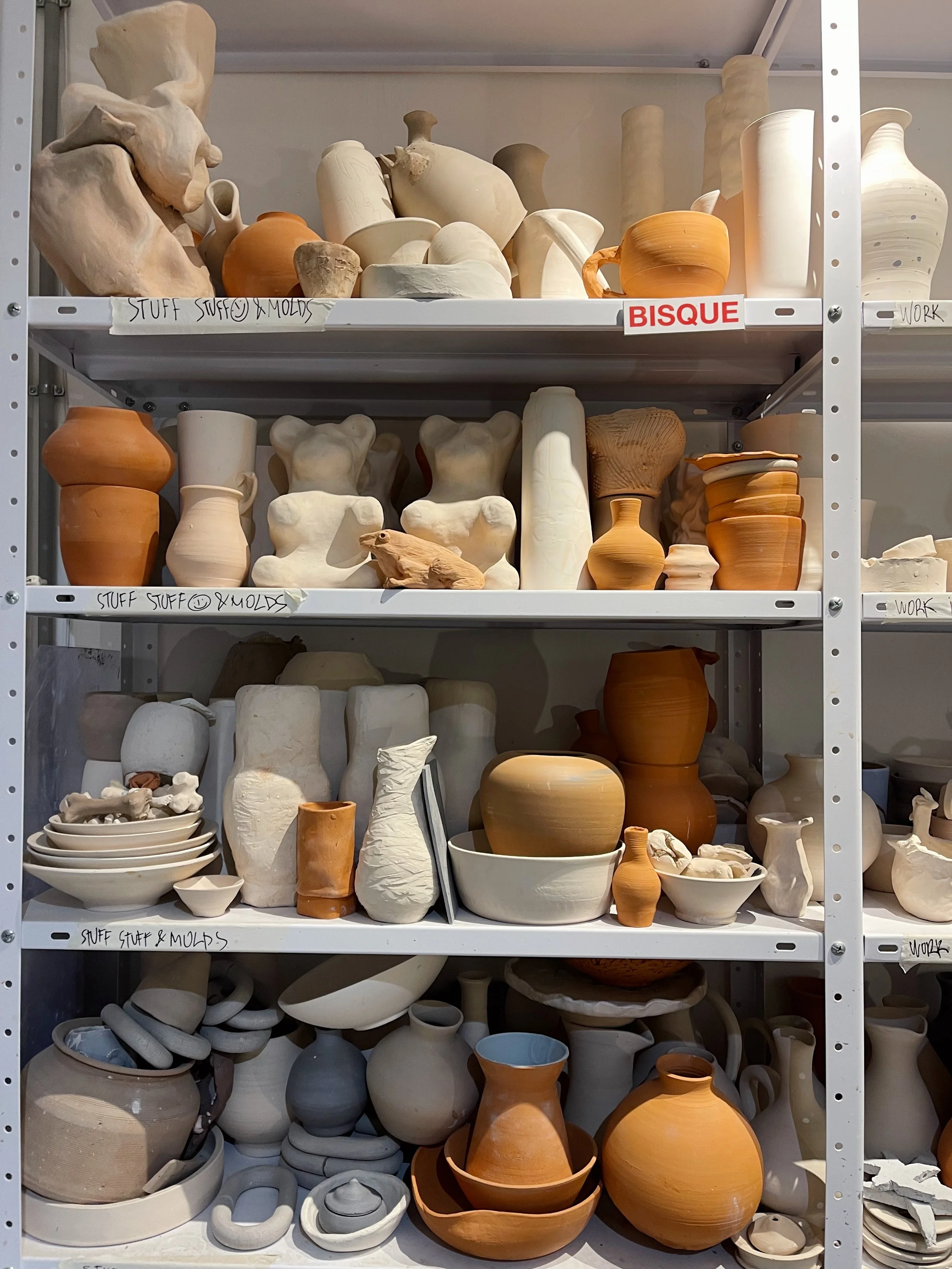 Shelves filled with various pottery, including vases, bowls, and mugs in shades of white, beige, and terracotta, labeled with categories like "Stuff Stuff @ Molds" and "Bisque."