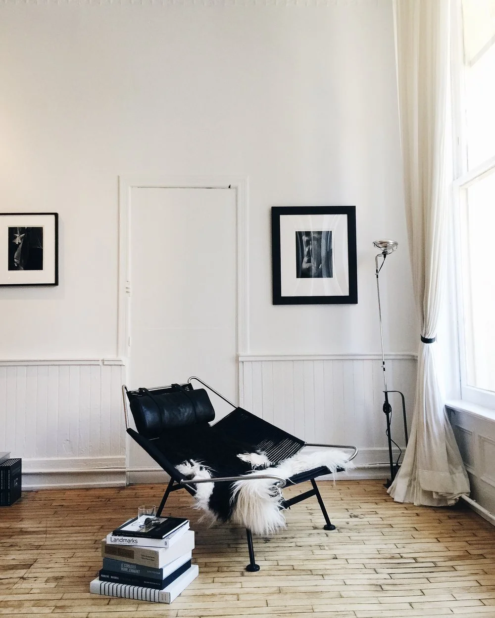 Minimalist living room with a black lounge chair draped with a black and white sheepskin rug, a stack of books, a glass of water, black and white framed artwork on the white walls, a tall floor lamp, and a window with cream curtains.
