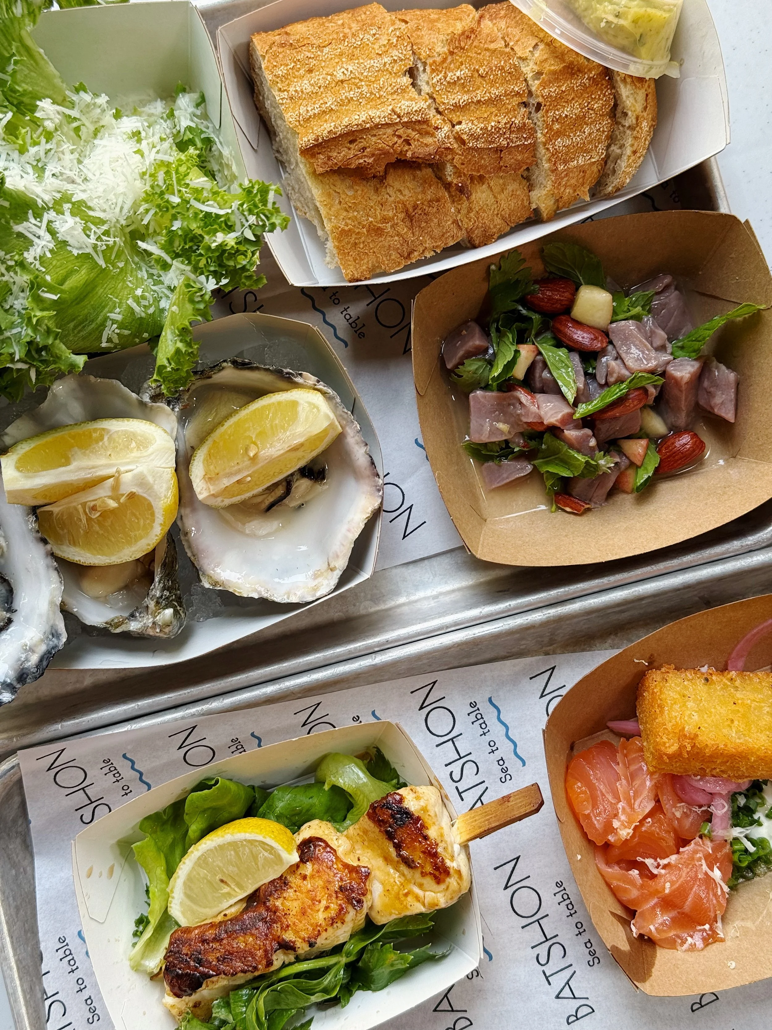 Assorted seafood and salad dishes including oysters with lemon wedges, a green leafy salad, toasted bread, a chicken skewer with lemon, a salmon and capers dish with crostini, and a salad with mixed greens, wild game meat, almonds, and a small contai