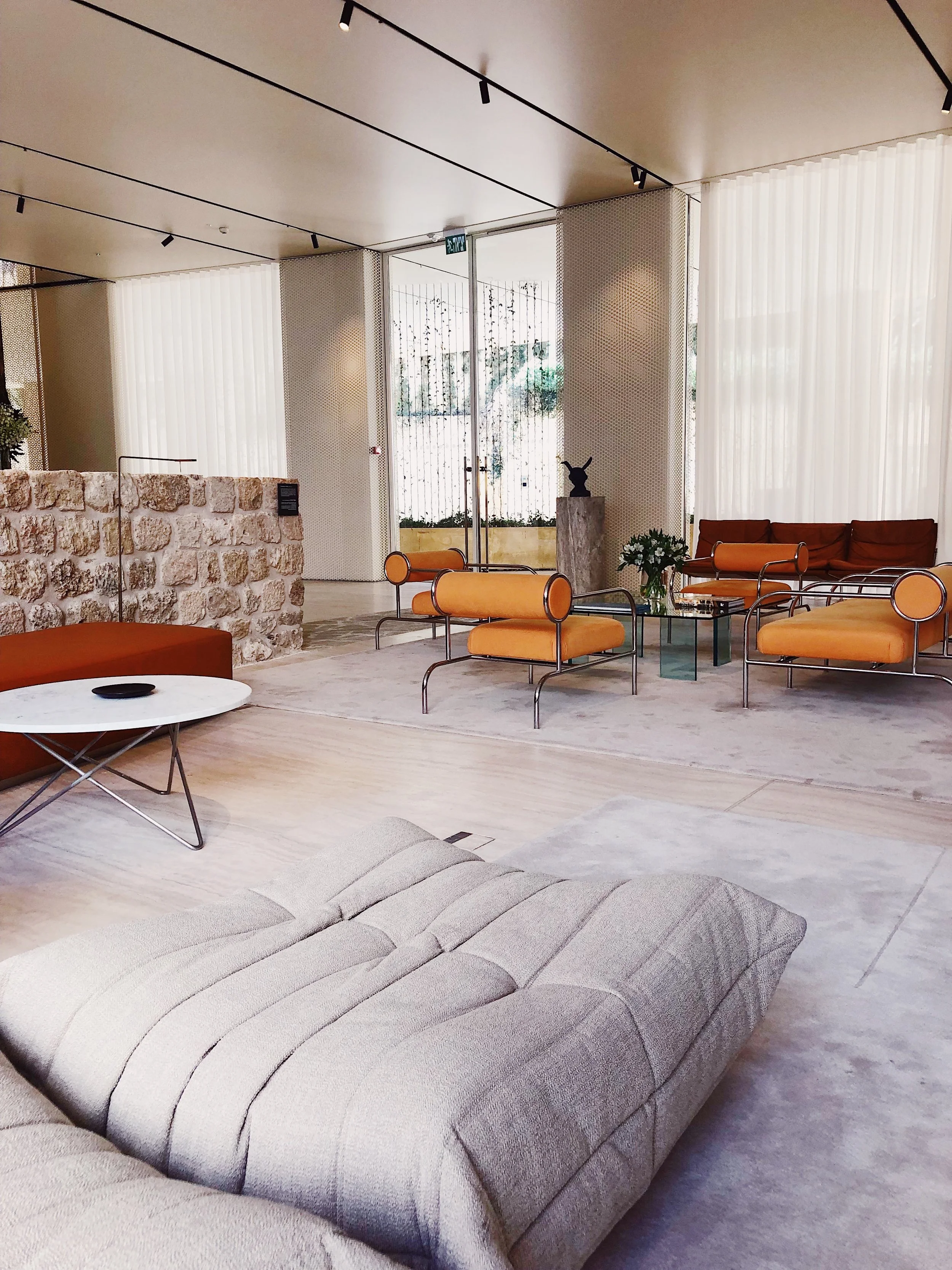 Modern hotel lobby with beige sectional sofa, orange chairs, glass coffee table, potted plant, stone wall, large windows with white curtains, and decorative sculpture.