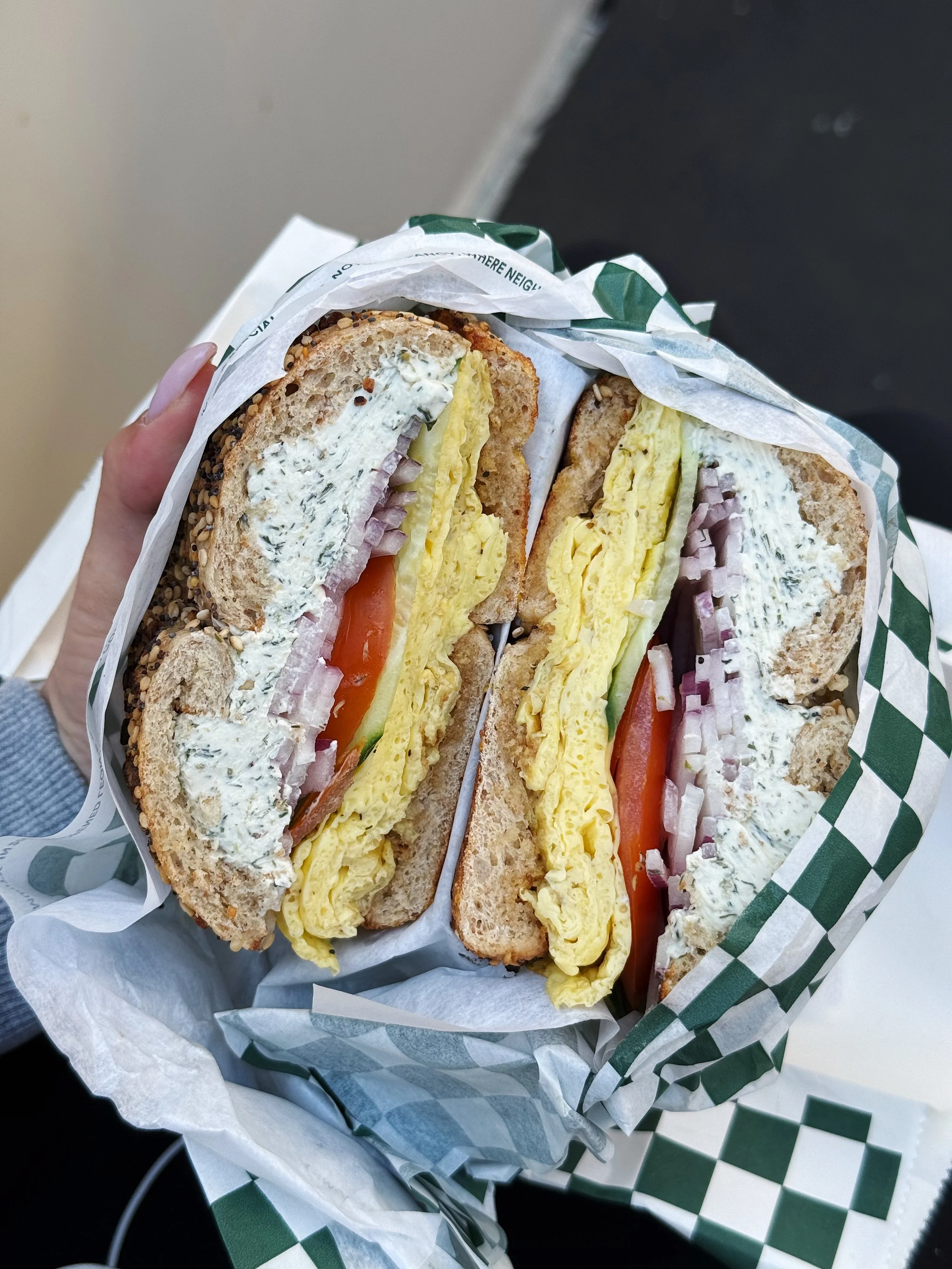 A sandwich cut in half showing layers of egg, tomato, cucumber, onion, and creamy spread on multigrain bread, wrapped in checkered paper.
