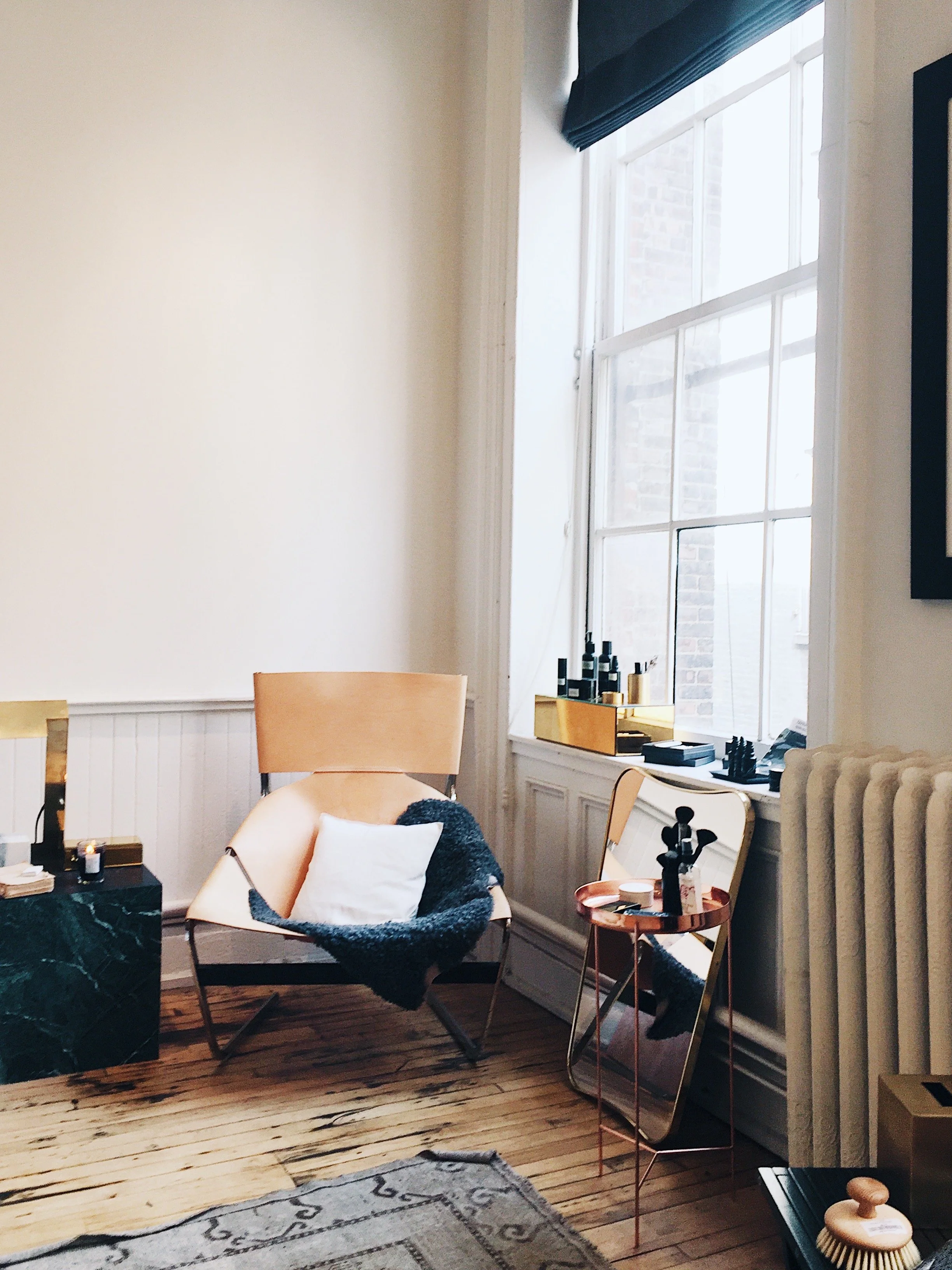 A cozy corner of a room with a beige armchair, a white pillow, and a black fuzzy throw blanket. There is a small copper side table with a black figurine, a mirror, a window with a black curtain, a radiator, and various bottles and decor items on the 