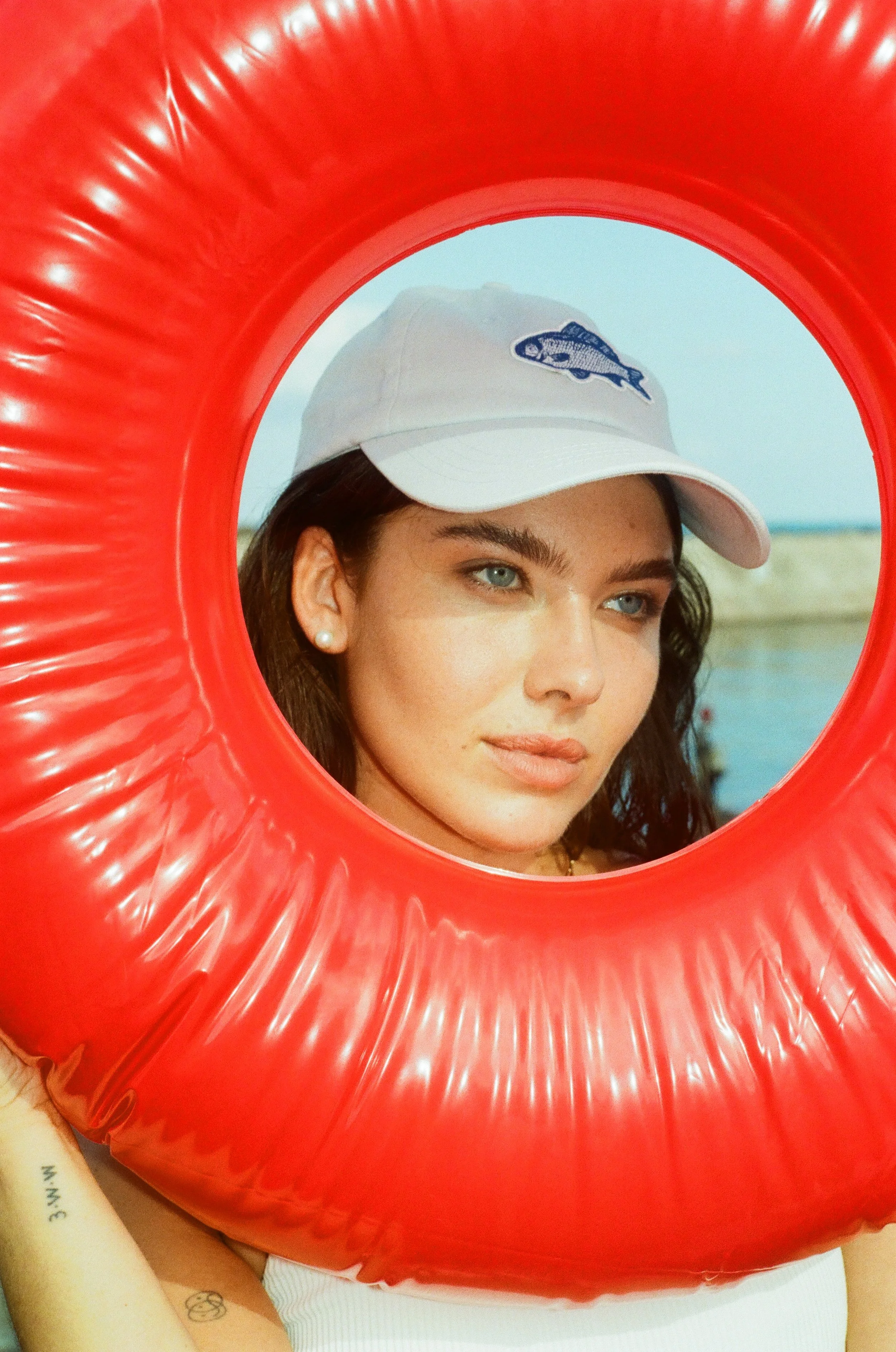 A young woman with blue eyes and dark hair wearing a white baseball cap with a fish logo, looking through a red inflatable tube at the beach or a body of water.