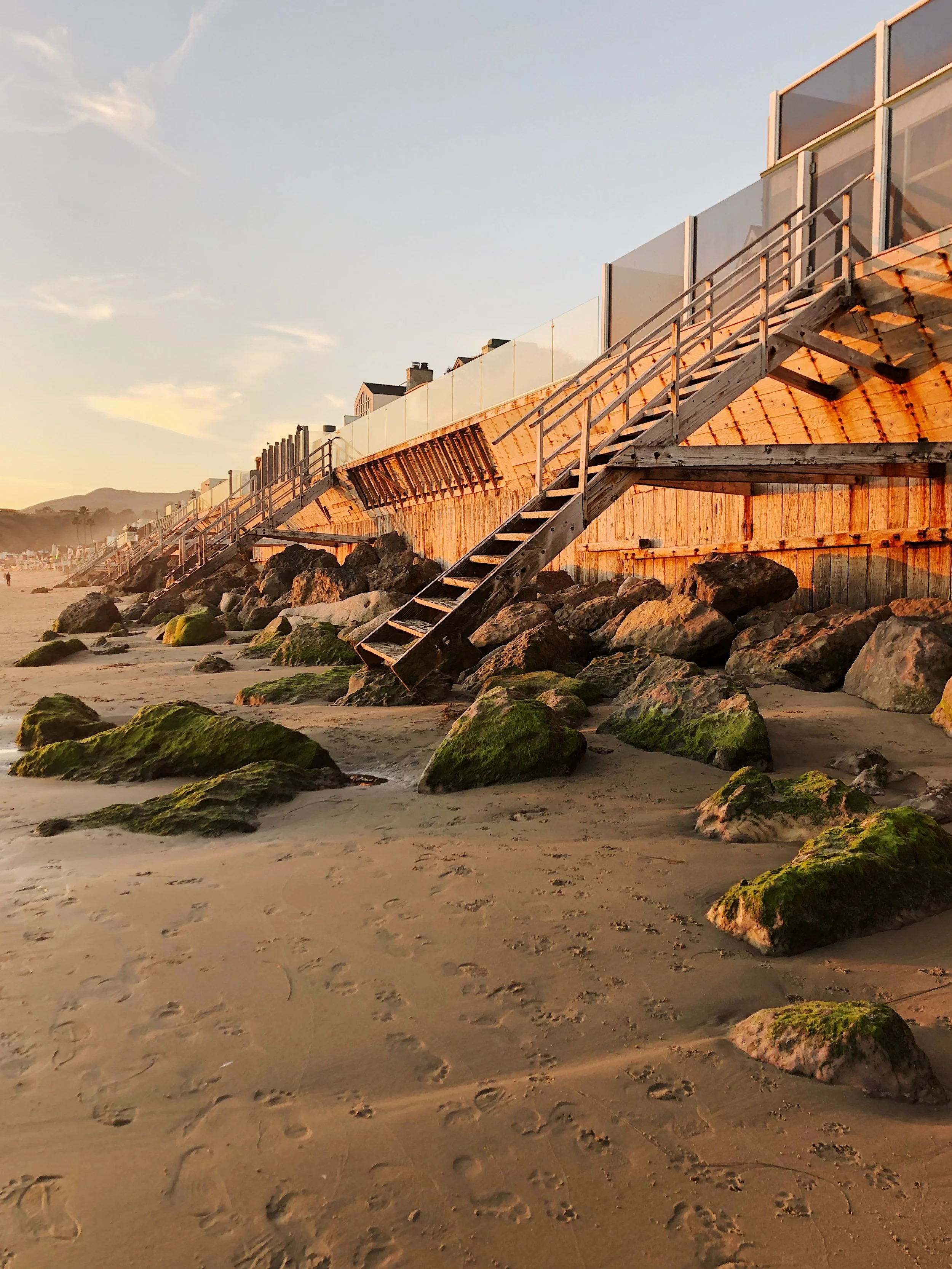 Sunset view of a beach with a wooden staircase leading from the sandy shore to a modern building with glass railings. Moss-covered rocks are scattered along the shoreline, and footprints are visible in the sand.