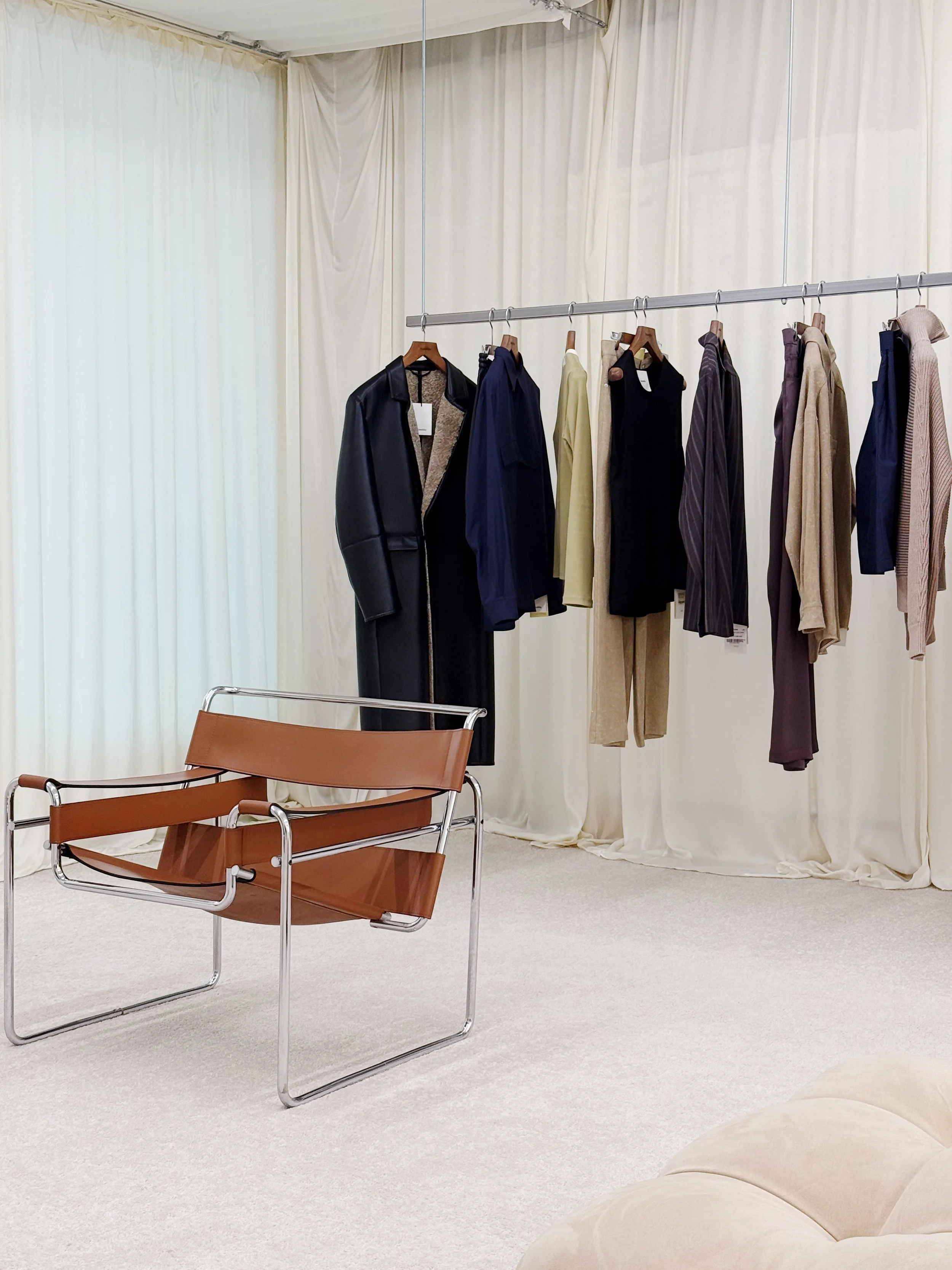 Clothing rack with various jackets and shirts, vintage brown leather and metal chair, cream curtains, and plush beige sofa in a minimalist room.