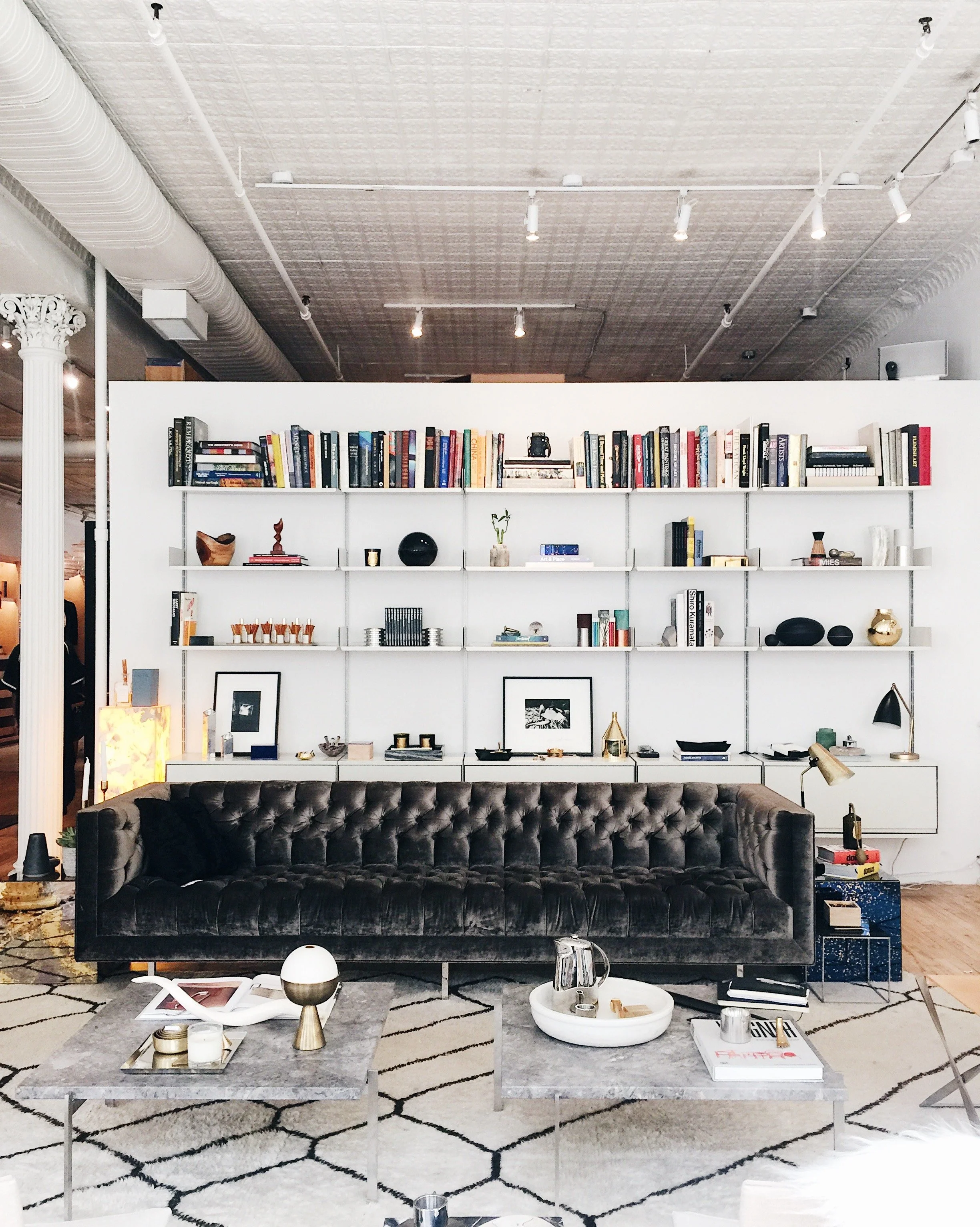 Living room with a black tufted sofa, two marble coffee tables, a white rug with black geometric pattern, and a large white bookshelf filled with books and decorative objects. There is a white wall with ceiling track lighting and an industrial ceilin
