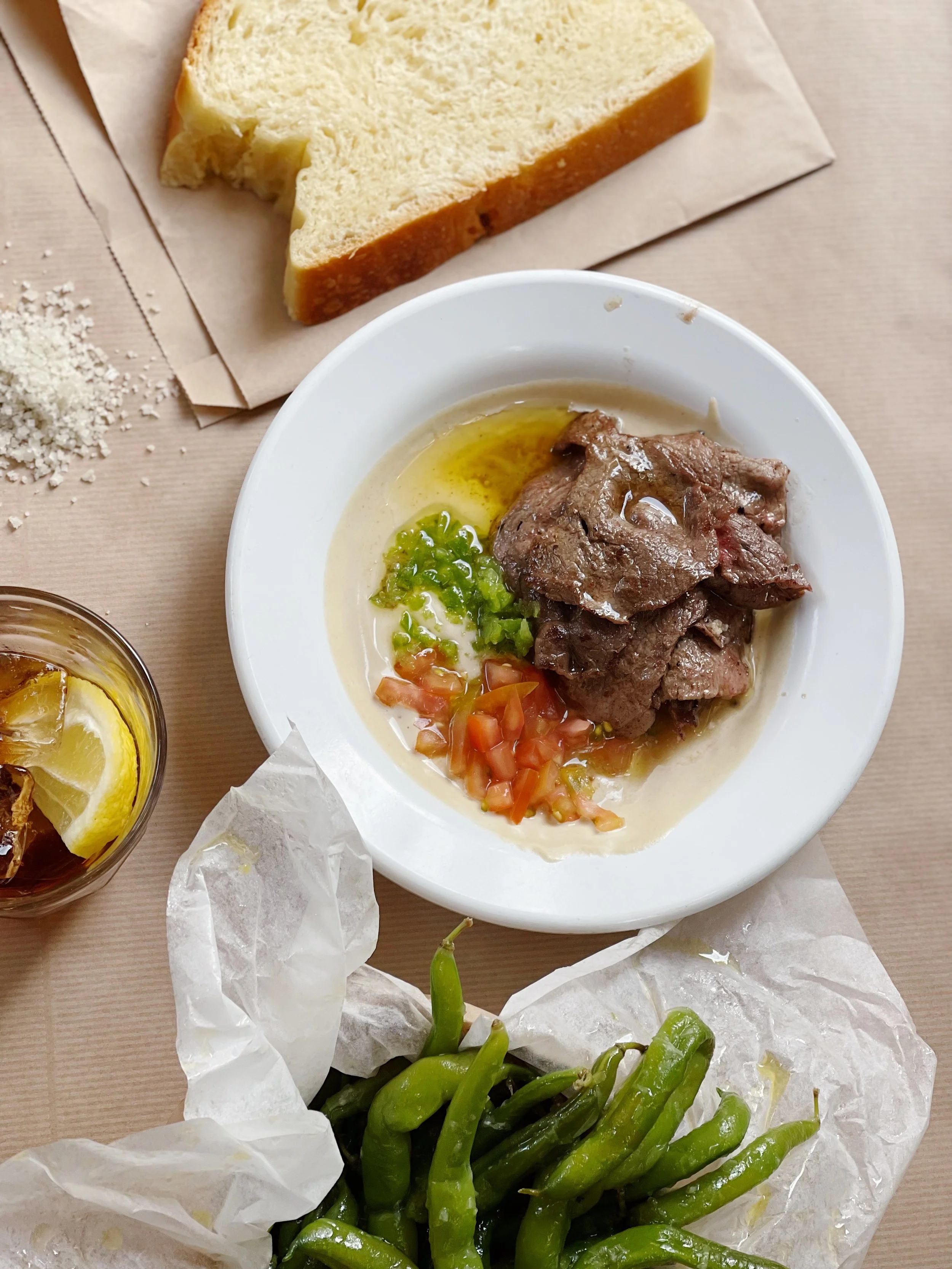 Plate of cooked meat with green and tomato garnish, slices of bread, green chili peppers, lemon ice tea, and a small pile of grated cheese on a beige table.
