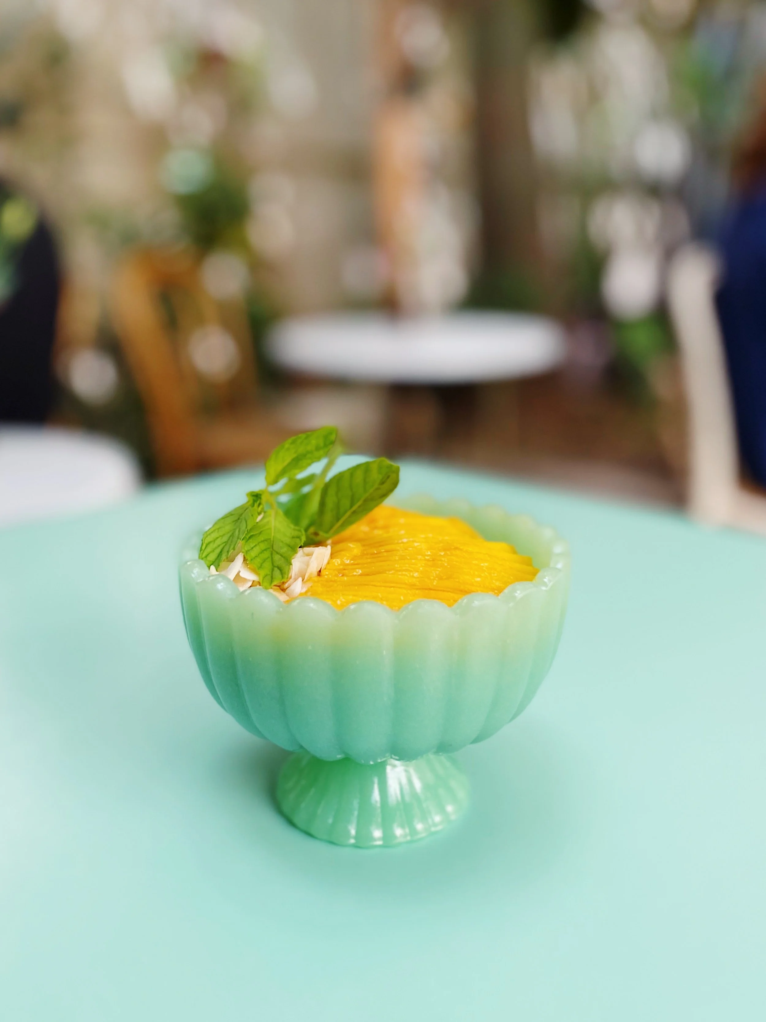 A bowl of yellow mango slices garnished with mint leaves and slivered almonds on a mint green table.