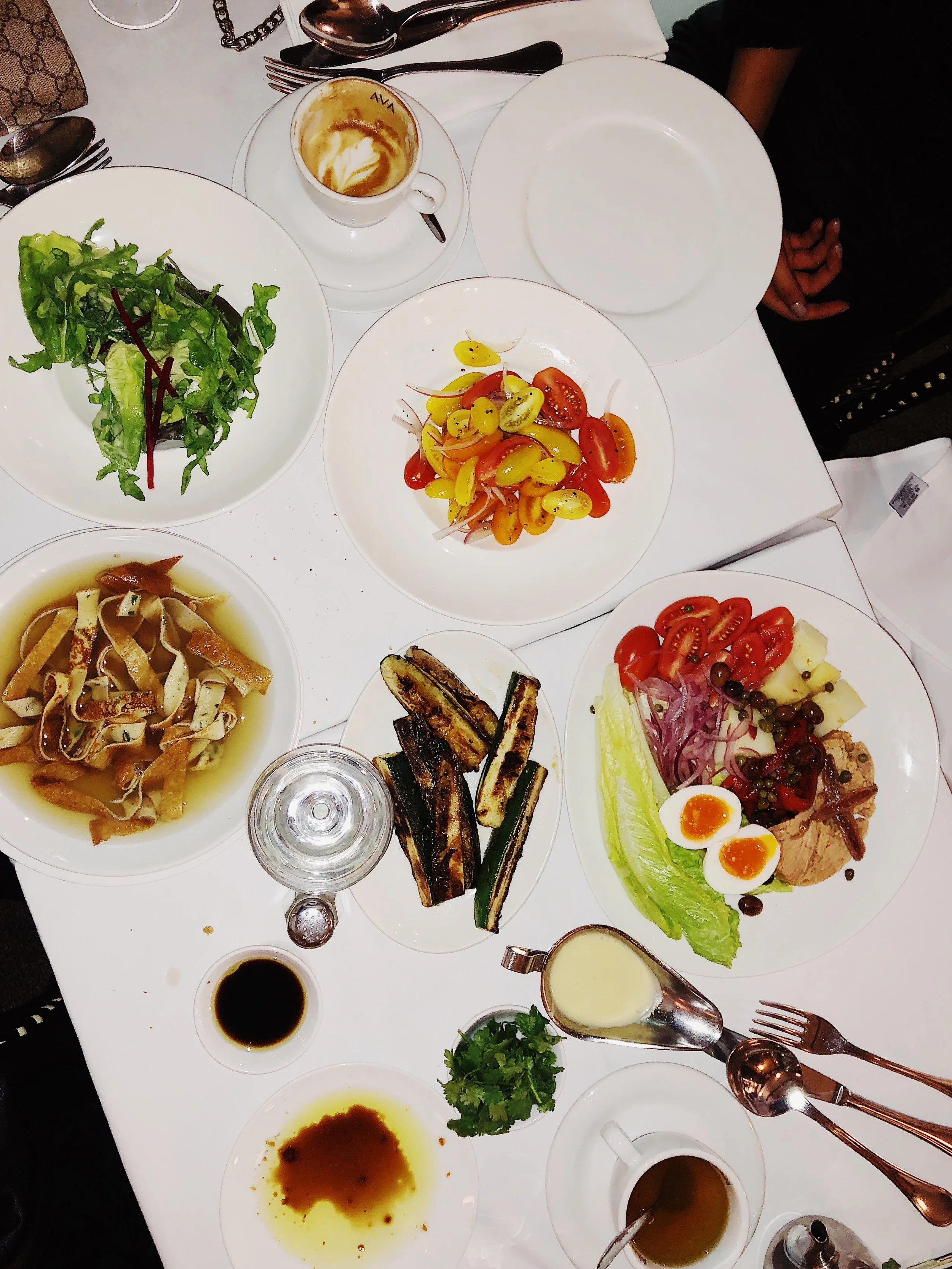 A table set with various dishes including a green salad, cherry tomatoes, grilled vegetables, pasta, and a plate with lettuce, tomatoes, eggs, and meat, with drinks and utensils.