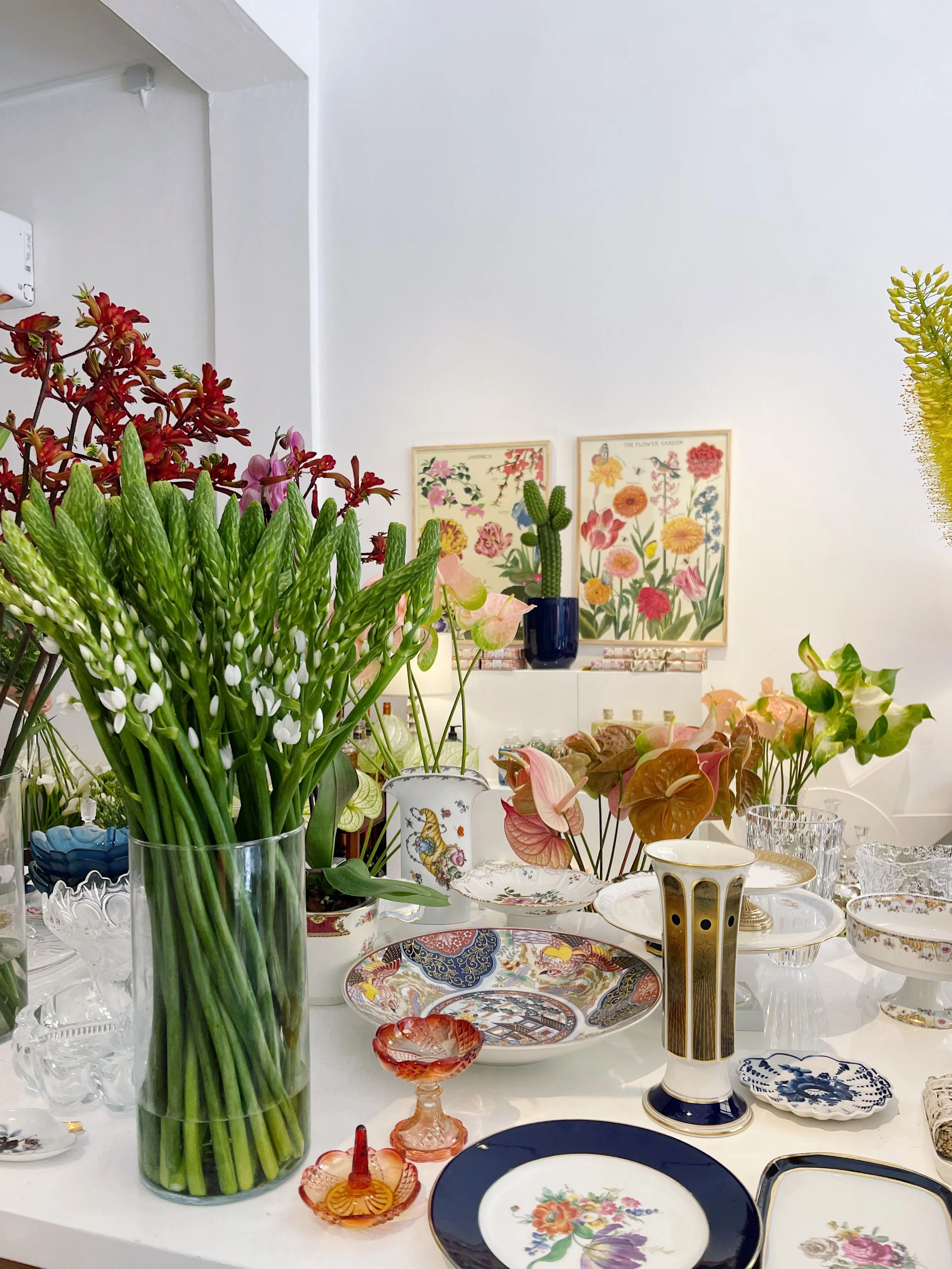 Display of various colorful decorative porcelain dishes, glassware, and a bouquet of flowers including lilies in a clear vase, arranged on a white table in a room with floral artwork on the wall.