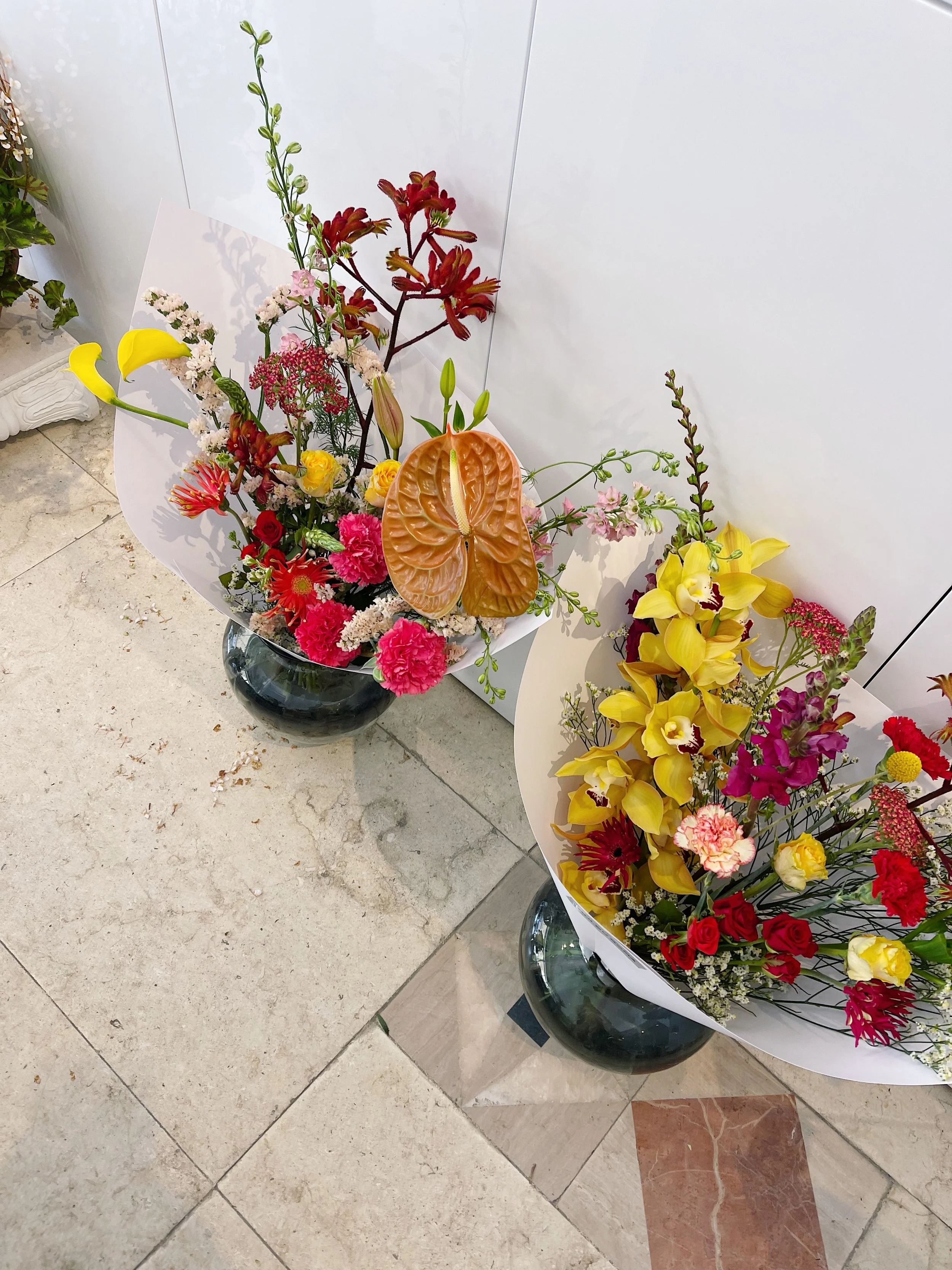 Two vibrant flower arrangements in black vases with a mix of yellow, pink, red, and white flowers, displayed on a tiled floor against a white wall.