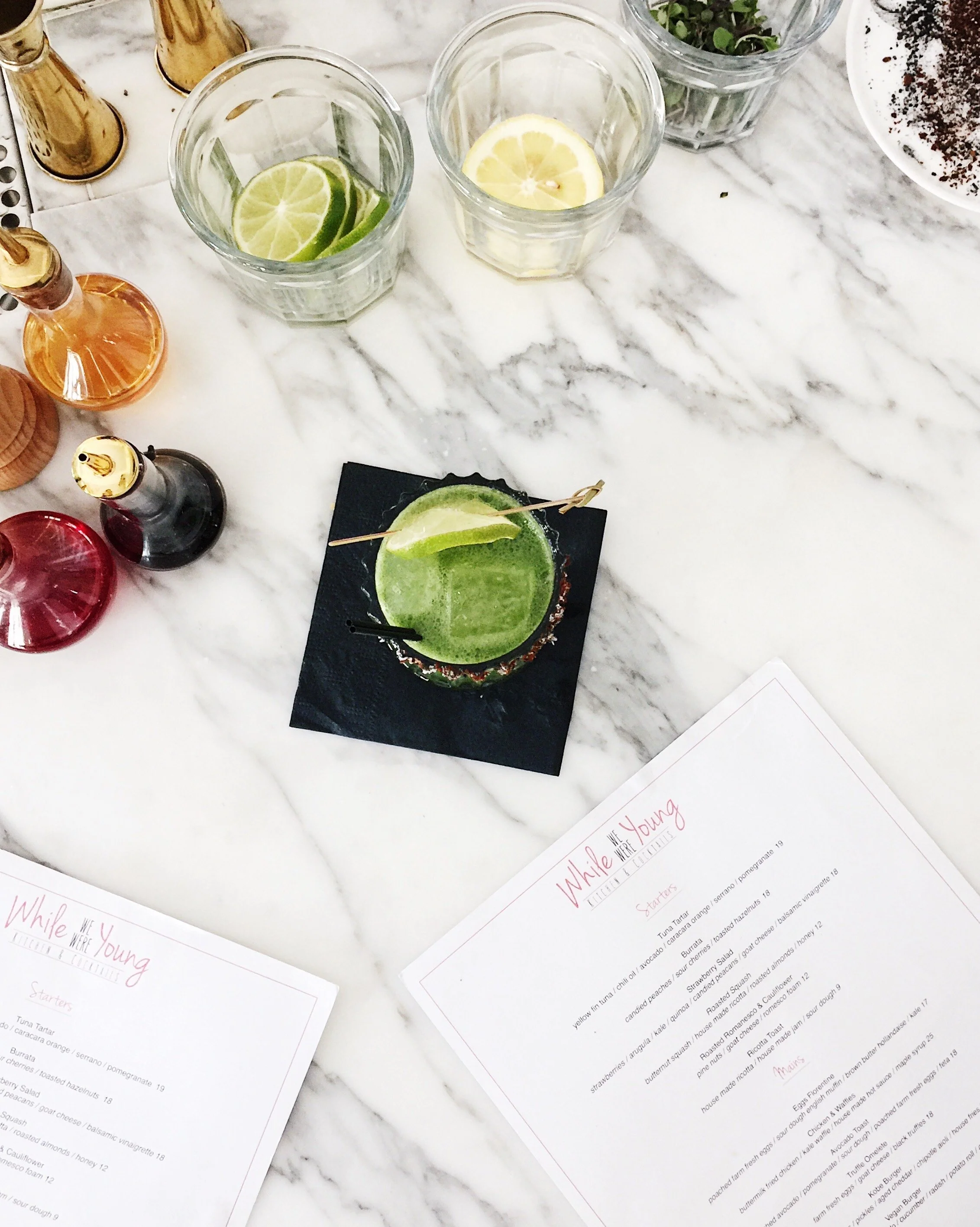 A marble countertop with three glasses containing lime and lemon slices, a cocktail with lime wedge and salt rim on a black coaster, condiment bottles, and two menus with pink text.