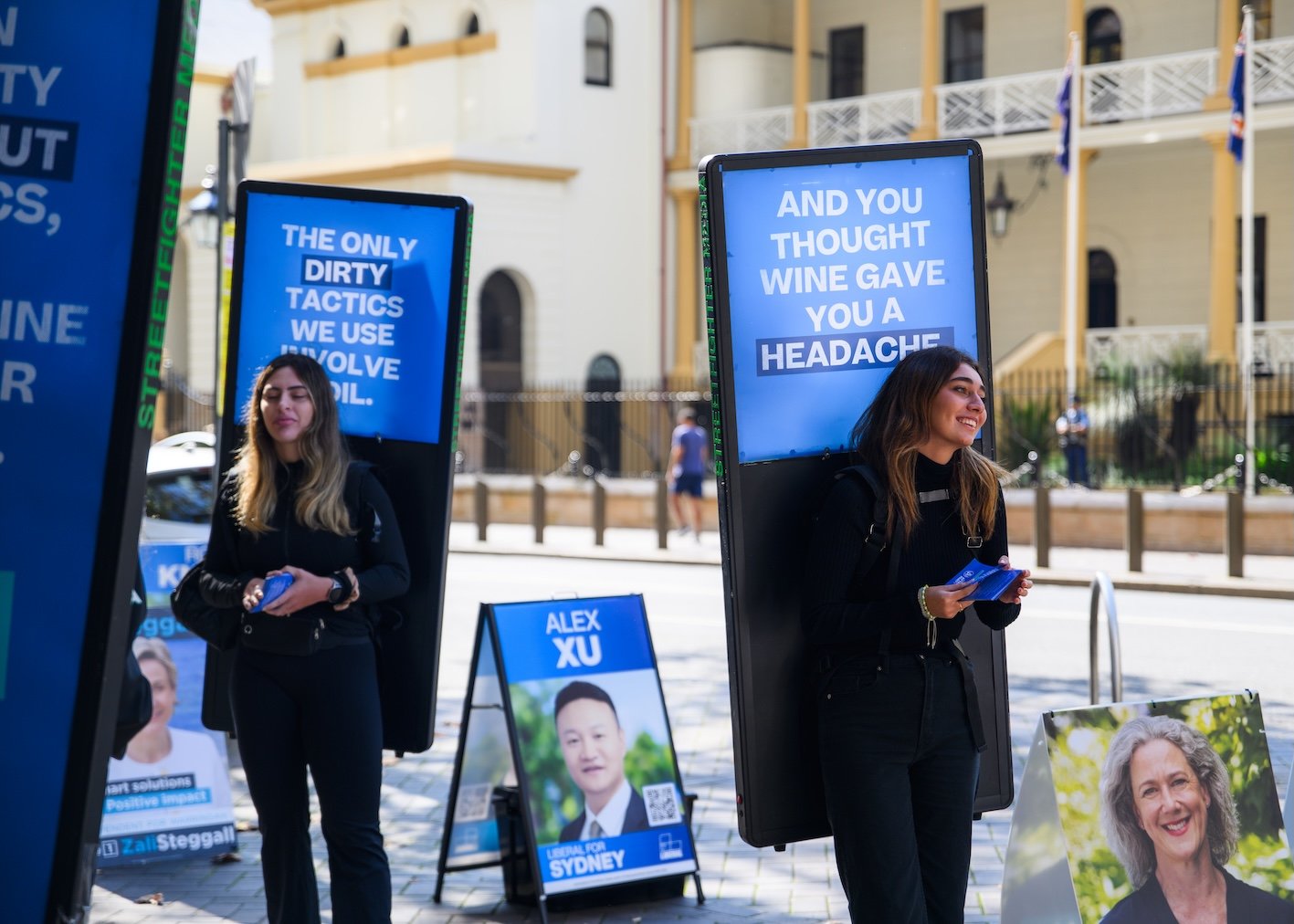 Two women standing outside at an election campaign event, with large blue campaign signs that have white text, one of which reads, 'The only dirty tactics we use involve oil,' and the other, 'And you thought wine gave you a headache.' They are smilin