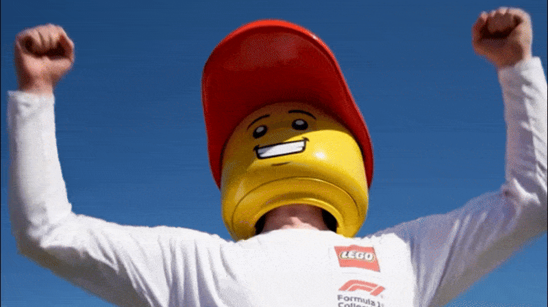 Person wearing a LEGO head mask with a red cap and a white long-sleeve shirt, raising fists in celebration against a blue sky.