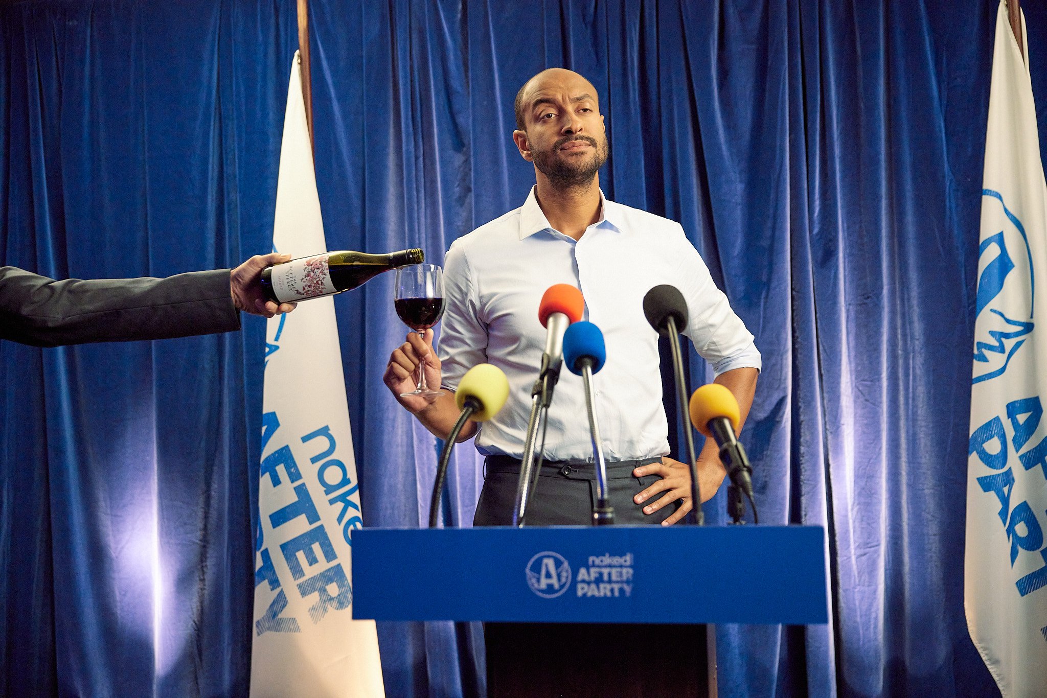 Man at podium with blue curtains and flags, holding a glass of red wine, while someone pours wine from a bottle.